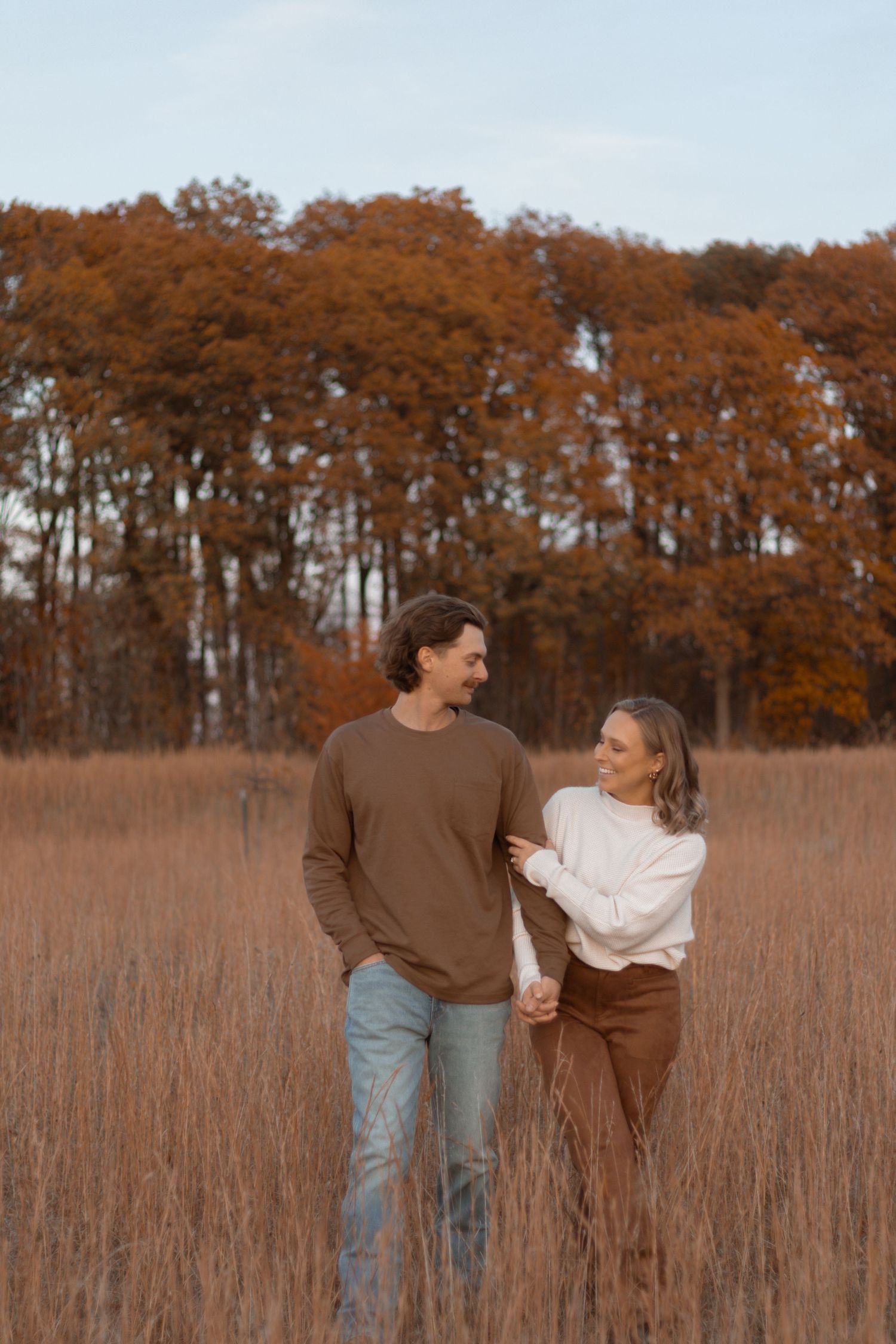 A couple embraces in a golden field of tall grass during autumn with rusty orange trees in the background.