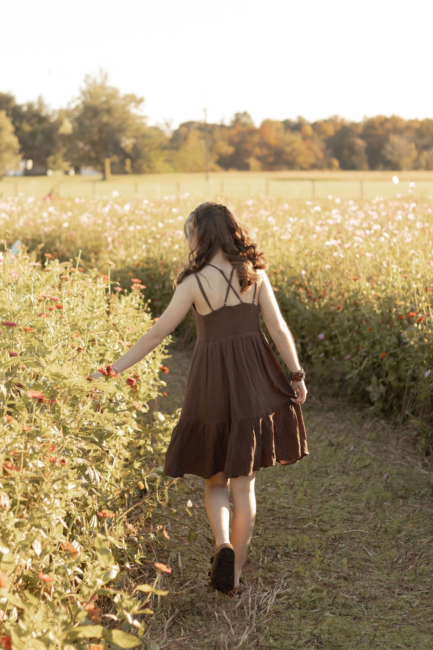 Woman in brown sundress walking along a wildflower field path in golden hour sunlight.