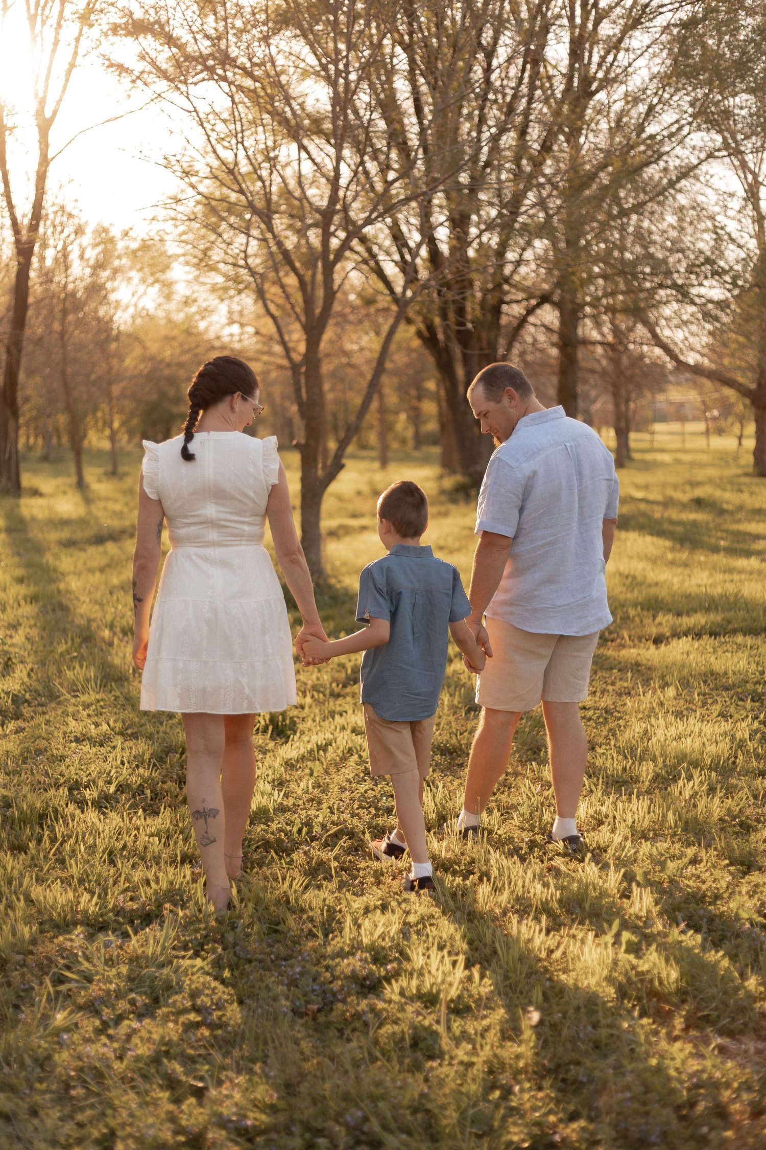 Family of three holding hands walking through a sunlit park at golden hour, viewed from behind.