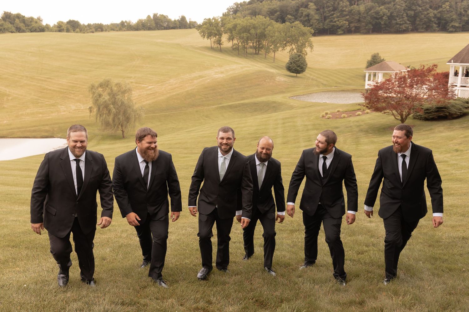 Six groomsmen in black suits walking together on a green lawn at an outdoor wedding venue.