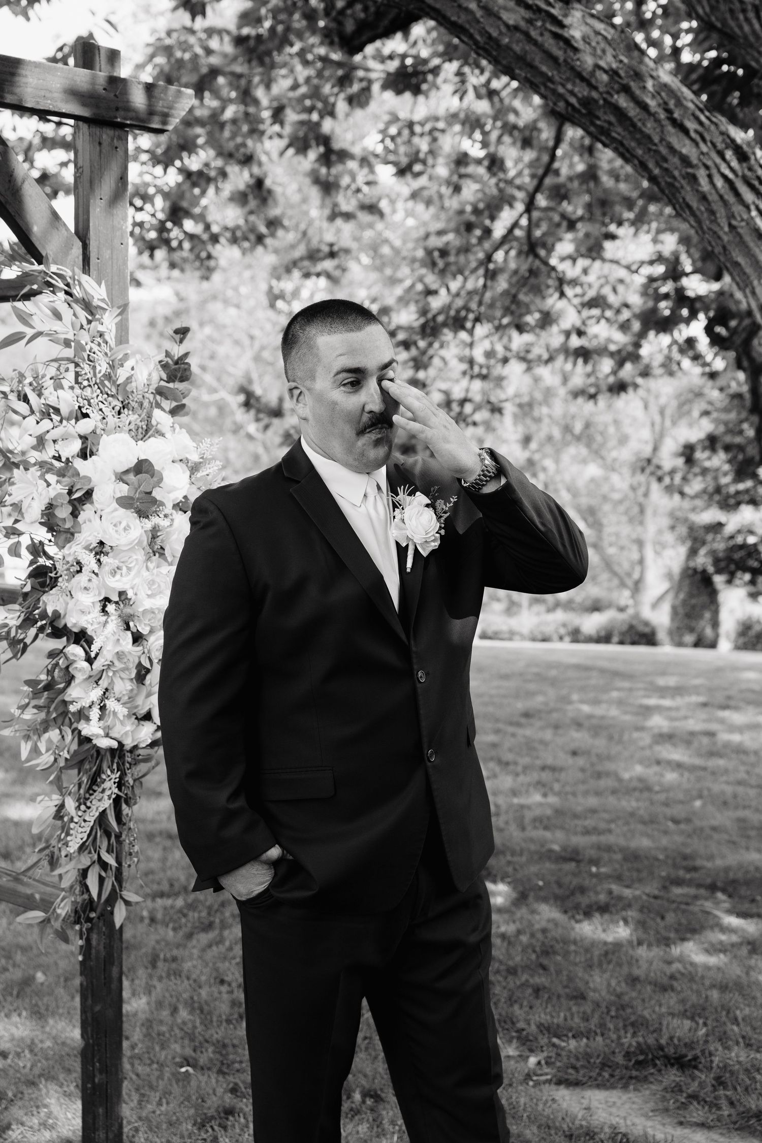 Groom in black suit wipes tear at outdoor wedding altar adorned with white floral arrangement, black and white photo.