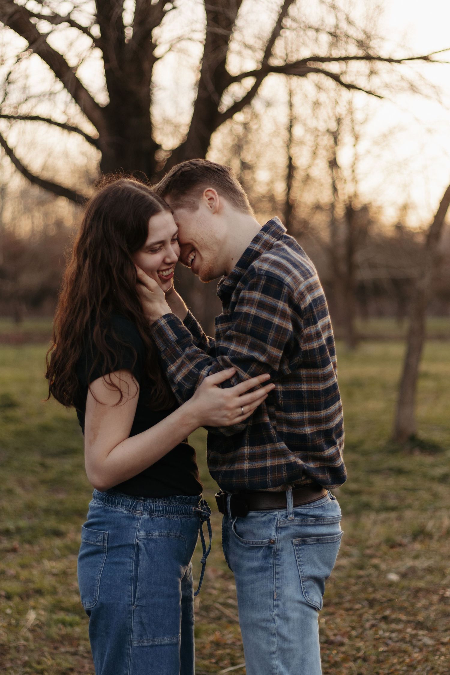 A couple laughs and embraces outdoors at sunset, wearing casual jeans and plaid, surrounded by bare trees.