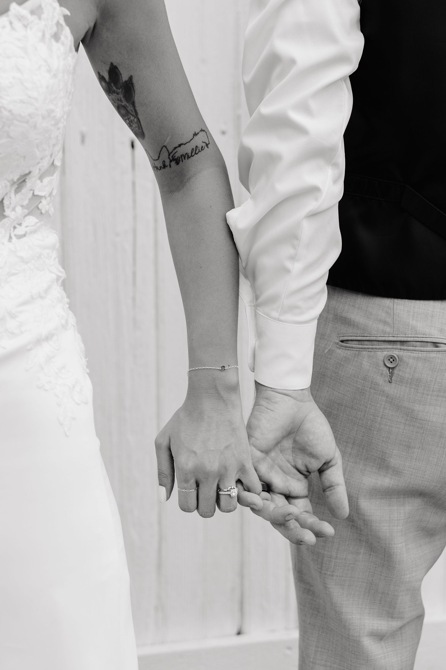 Black and white photo of a bride and groom holding hands, showing wedding rings and a floral tattoo on the bride's wrist.