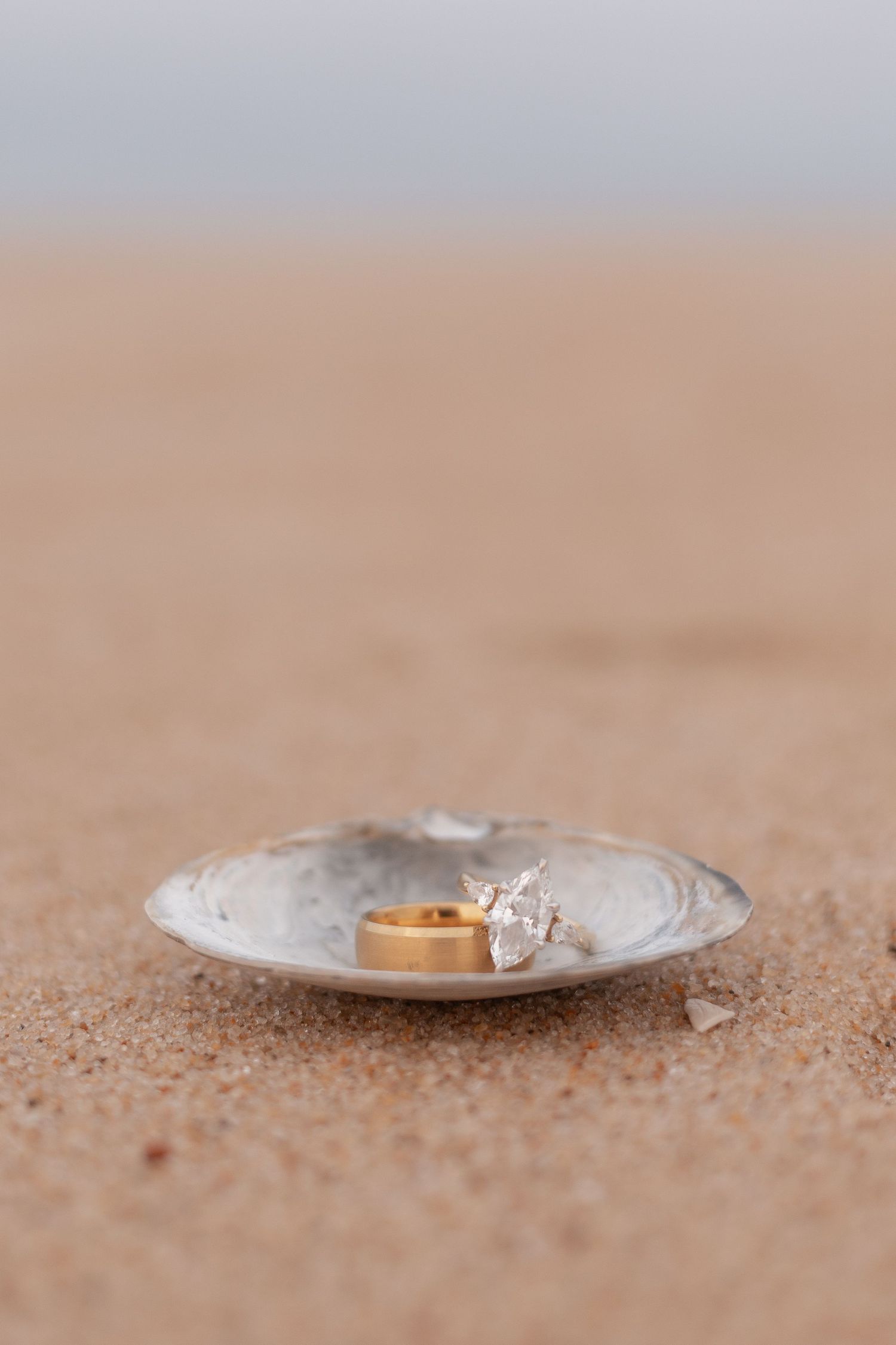 Gold wedding band and diamond engagement ring resting in a seashell on sandy beach.