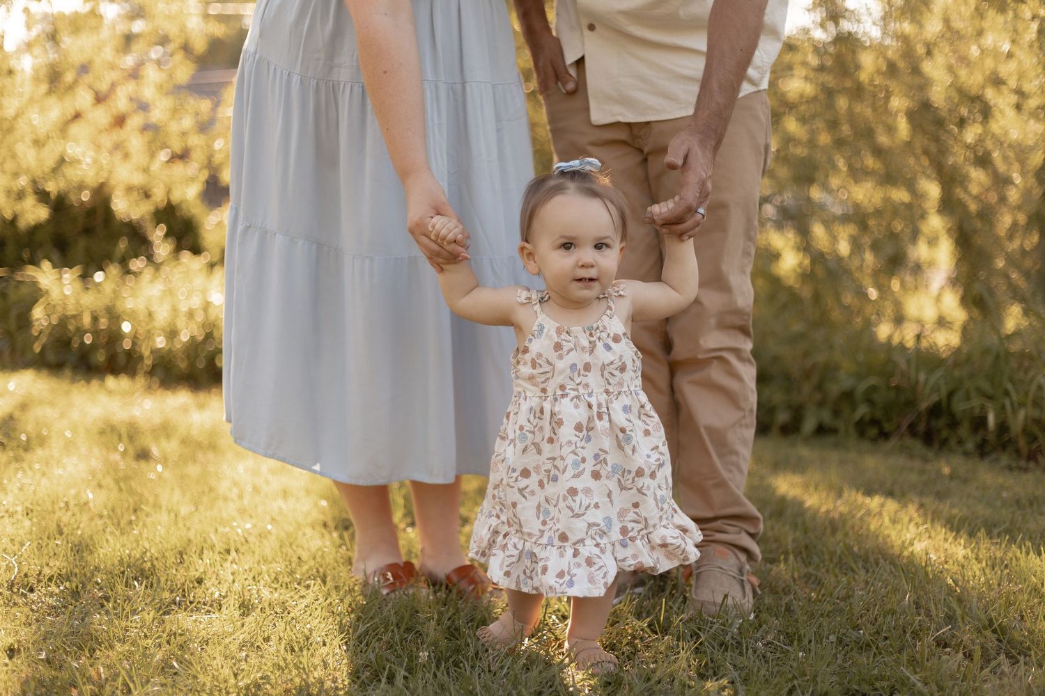 A toddler in a floral dress takes first steps holding parents hands in a sunny green garden.