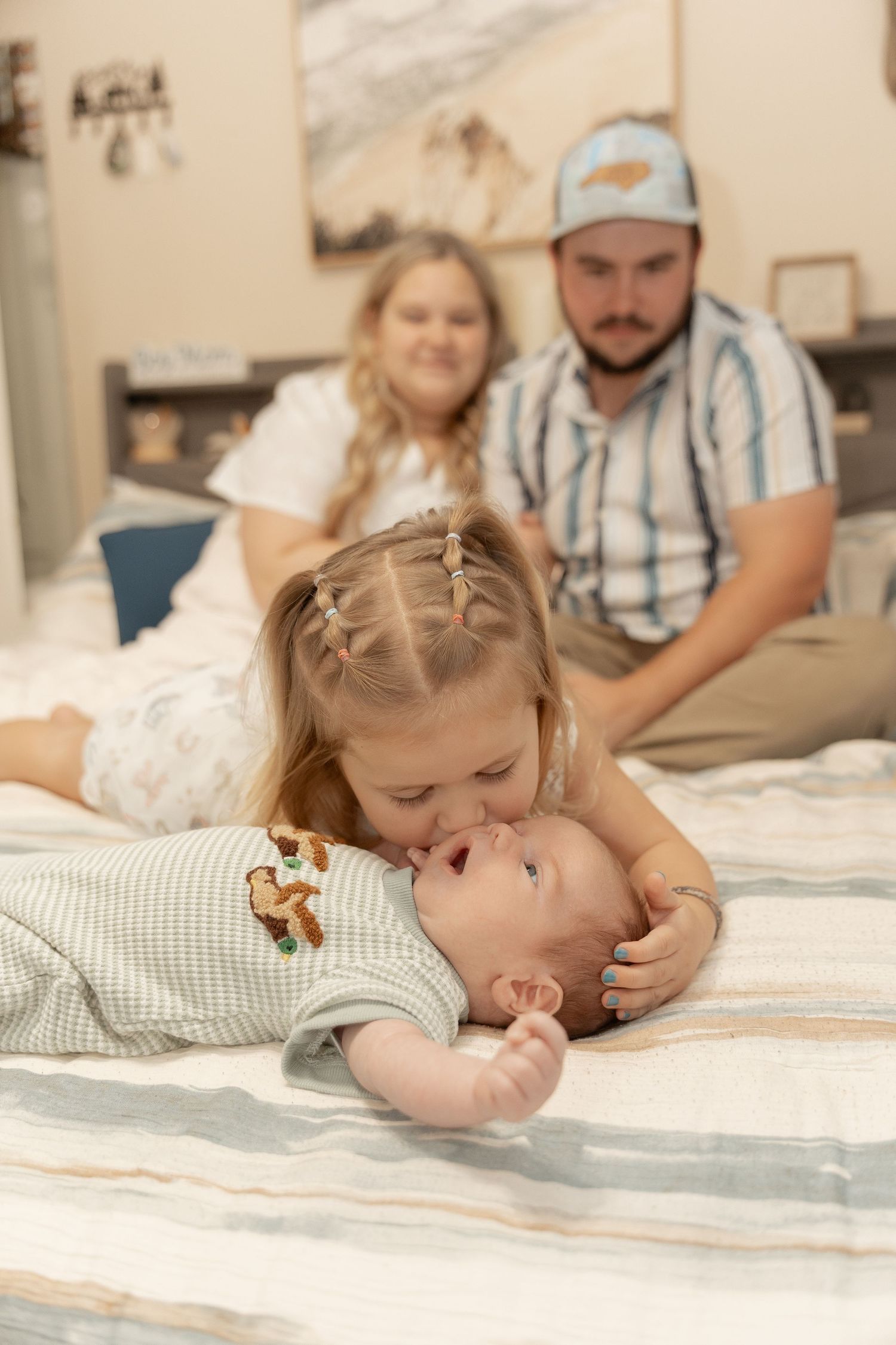 A toddler girl with braids kisses a newborn baby on a bed while parents smile warmly in the background.