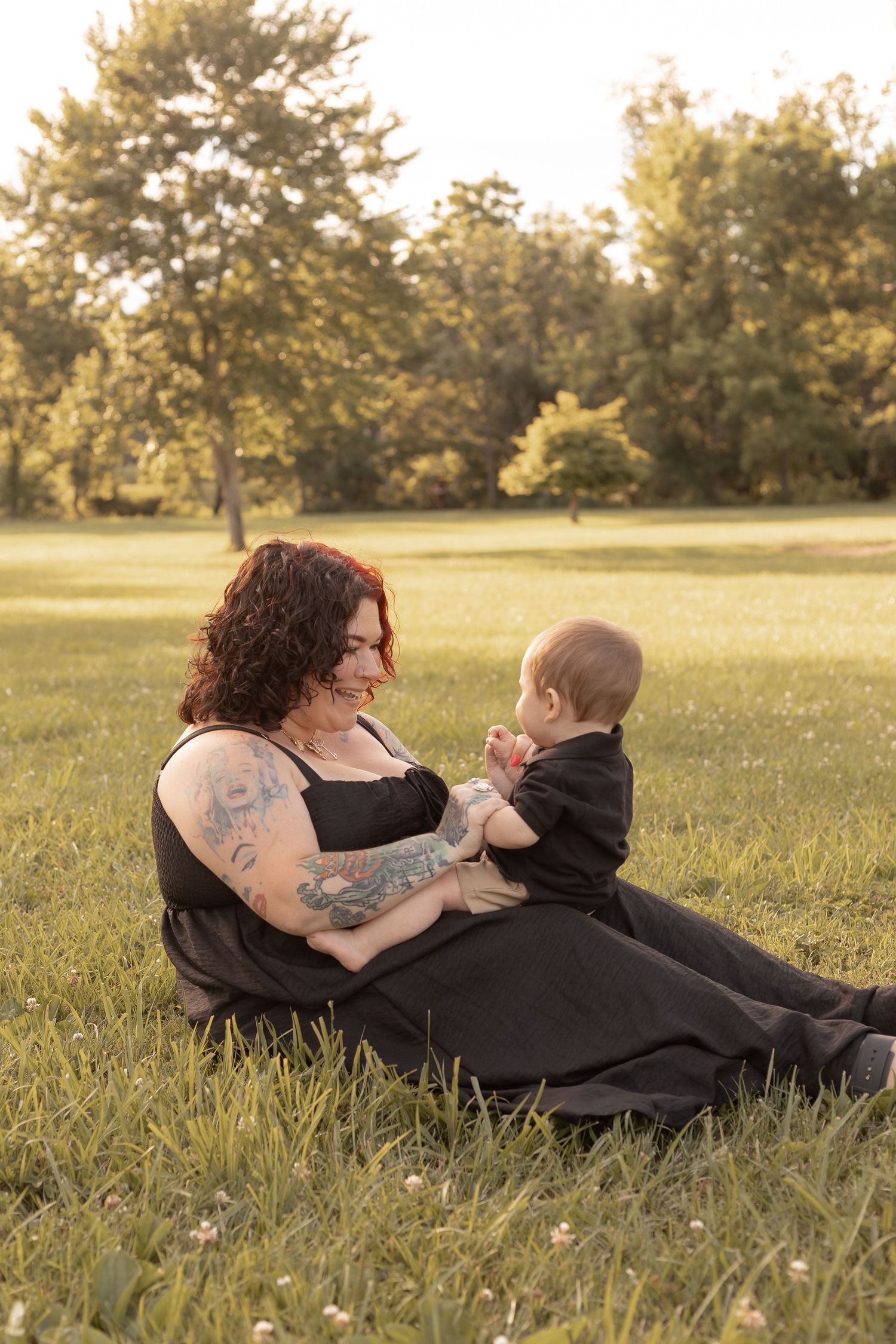 A tattooed mother sits on green grass holding her baby in a sunny park, both dressed in black.