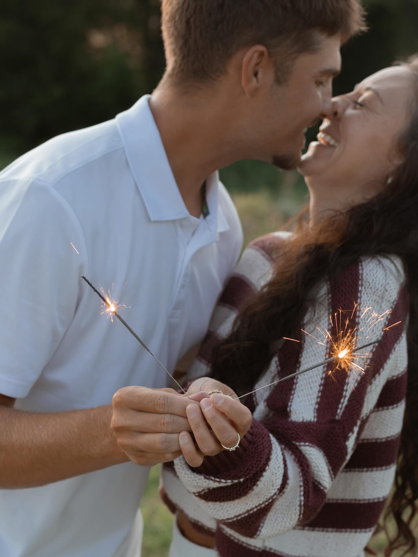 A couple shares an intimate moment while holding sparklers at dusk, dressed in white and burgundy sweater.