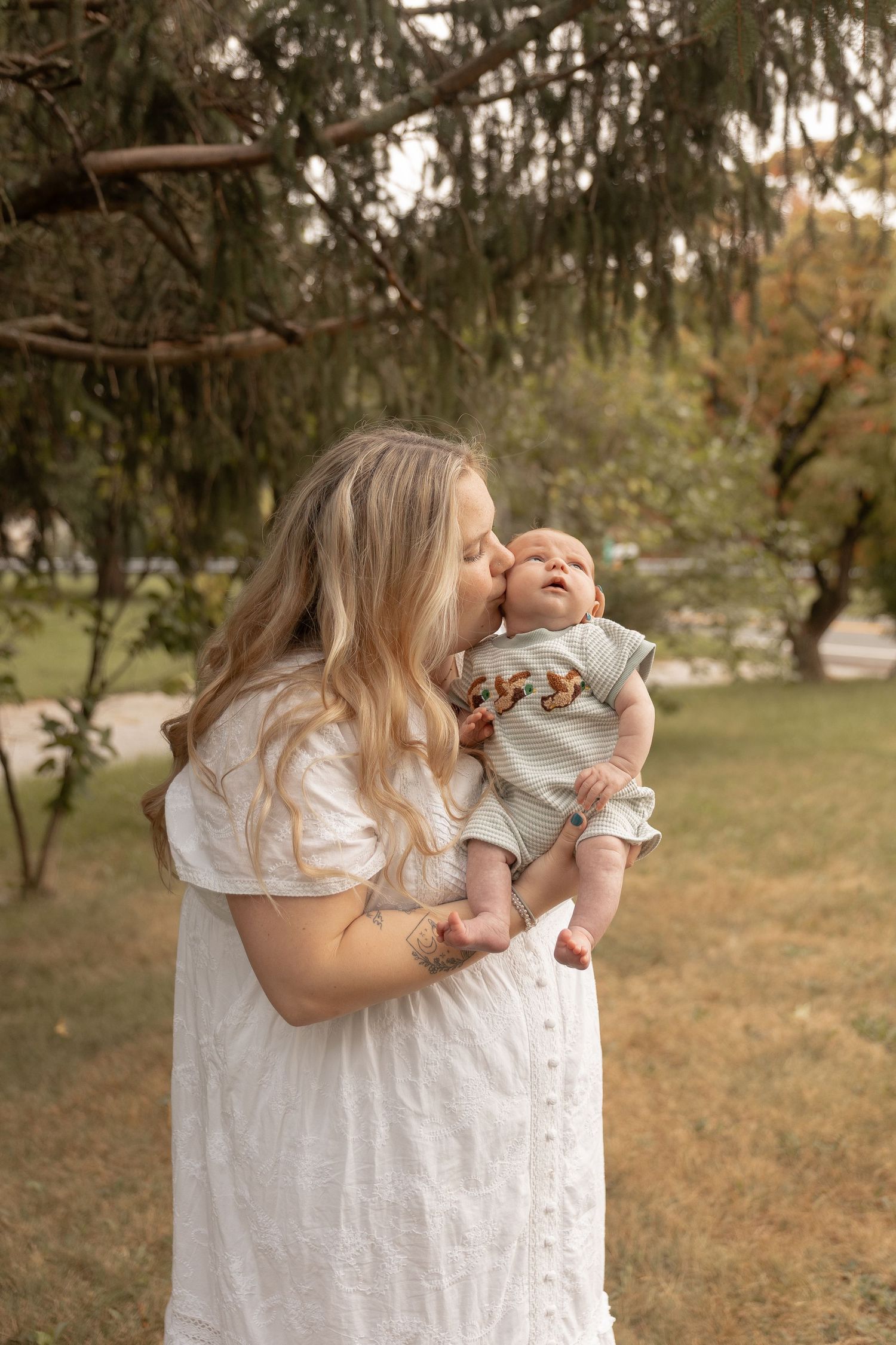A blonde woman in a white dress kisses her newborn baby outdoors in a lush green park setting.
