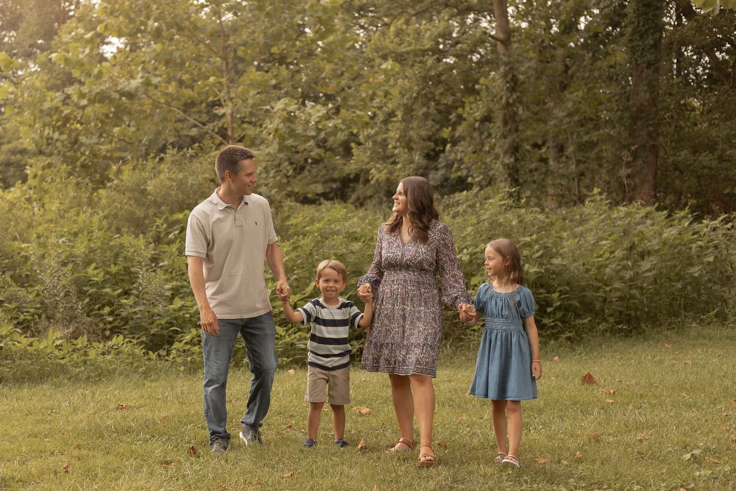 Family of four holding hands and walking in a lush green park during golden hour outdoor photo session.