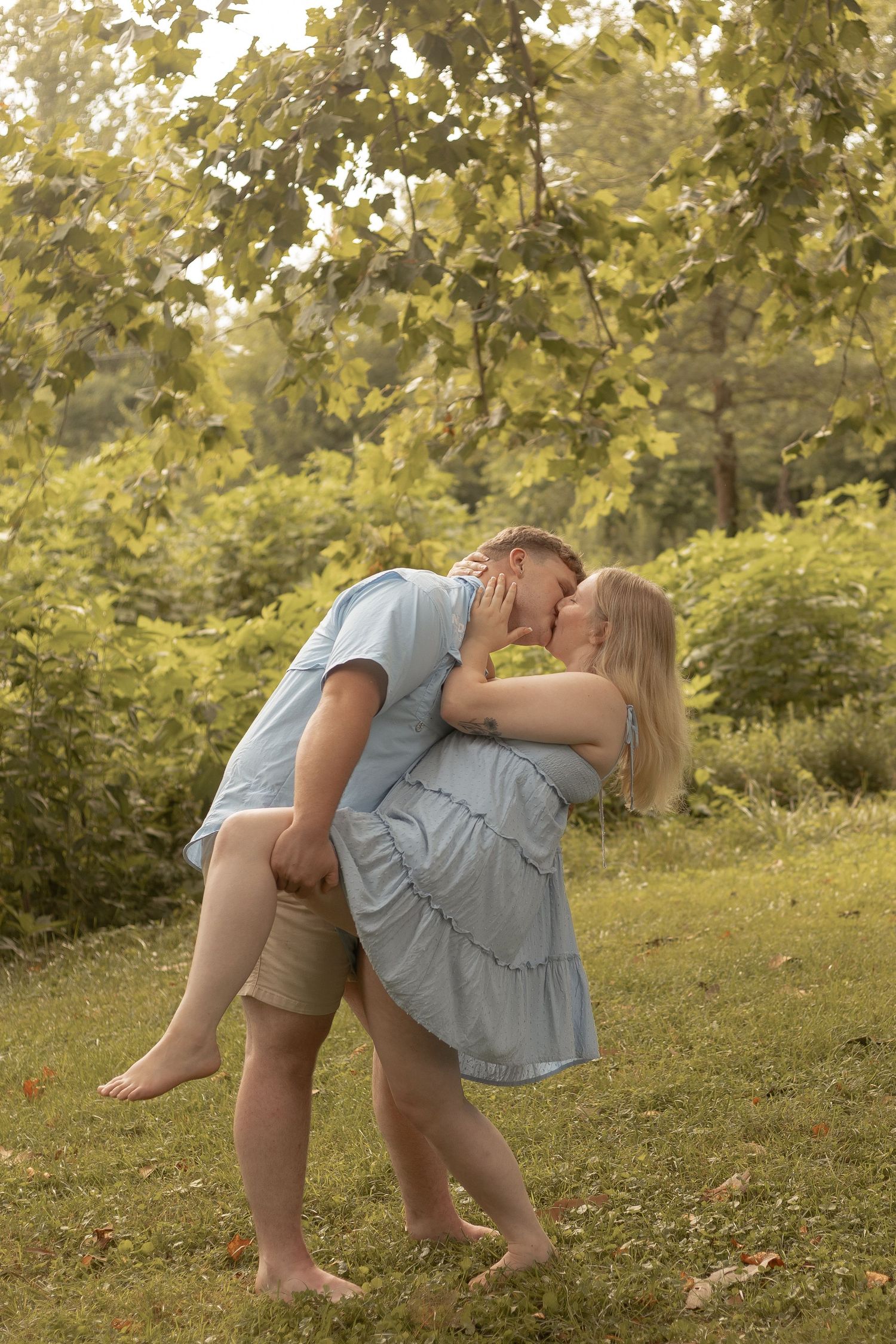A couple shares a romantic kiss while embracing outdoors in a lush green park setting.