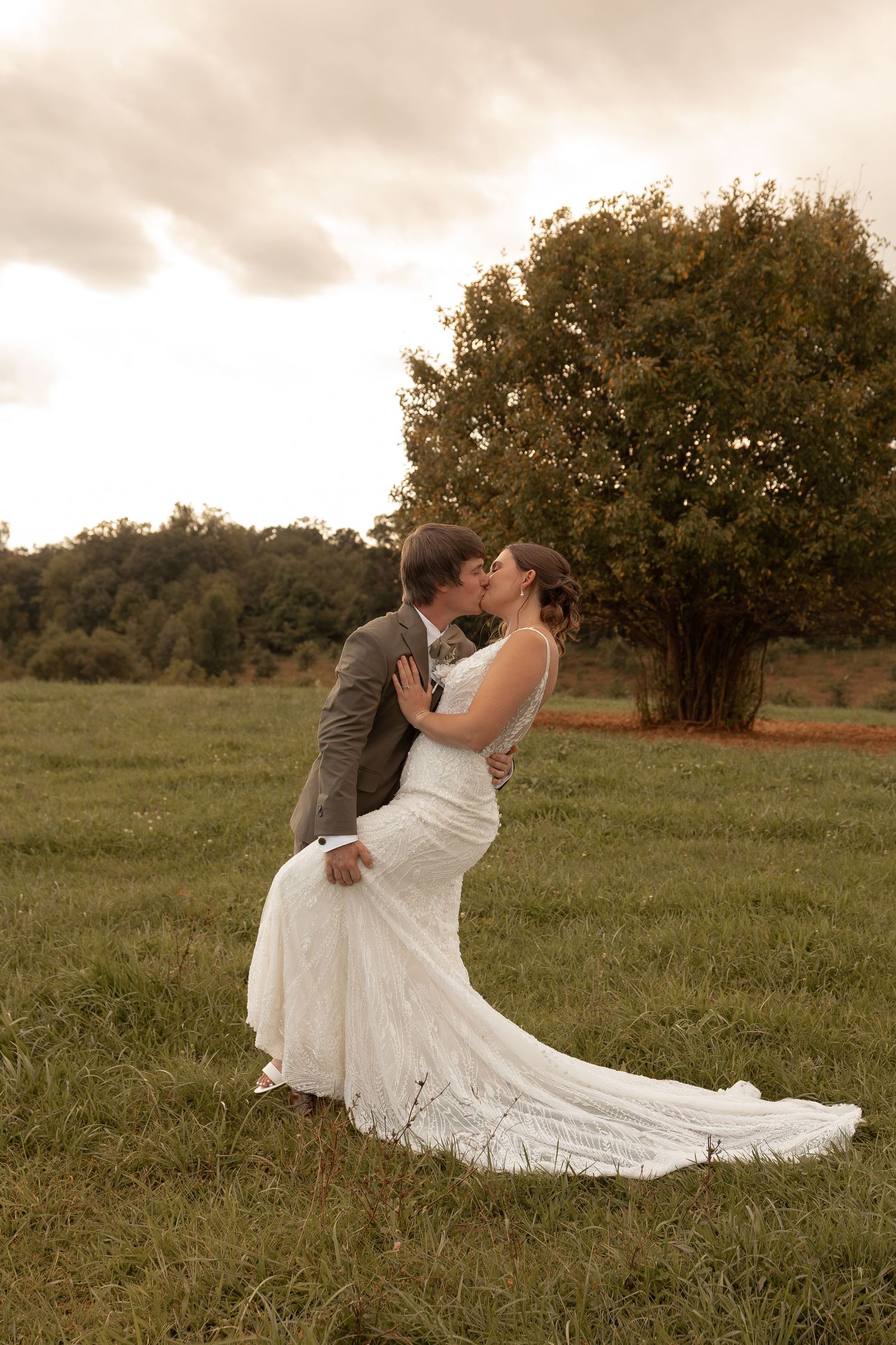 Bride and groom share a romantic kiss in a lush green field with a large tree backdrop on their wedding day.