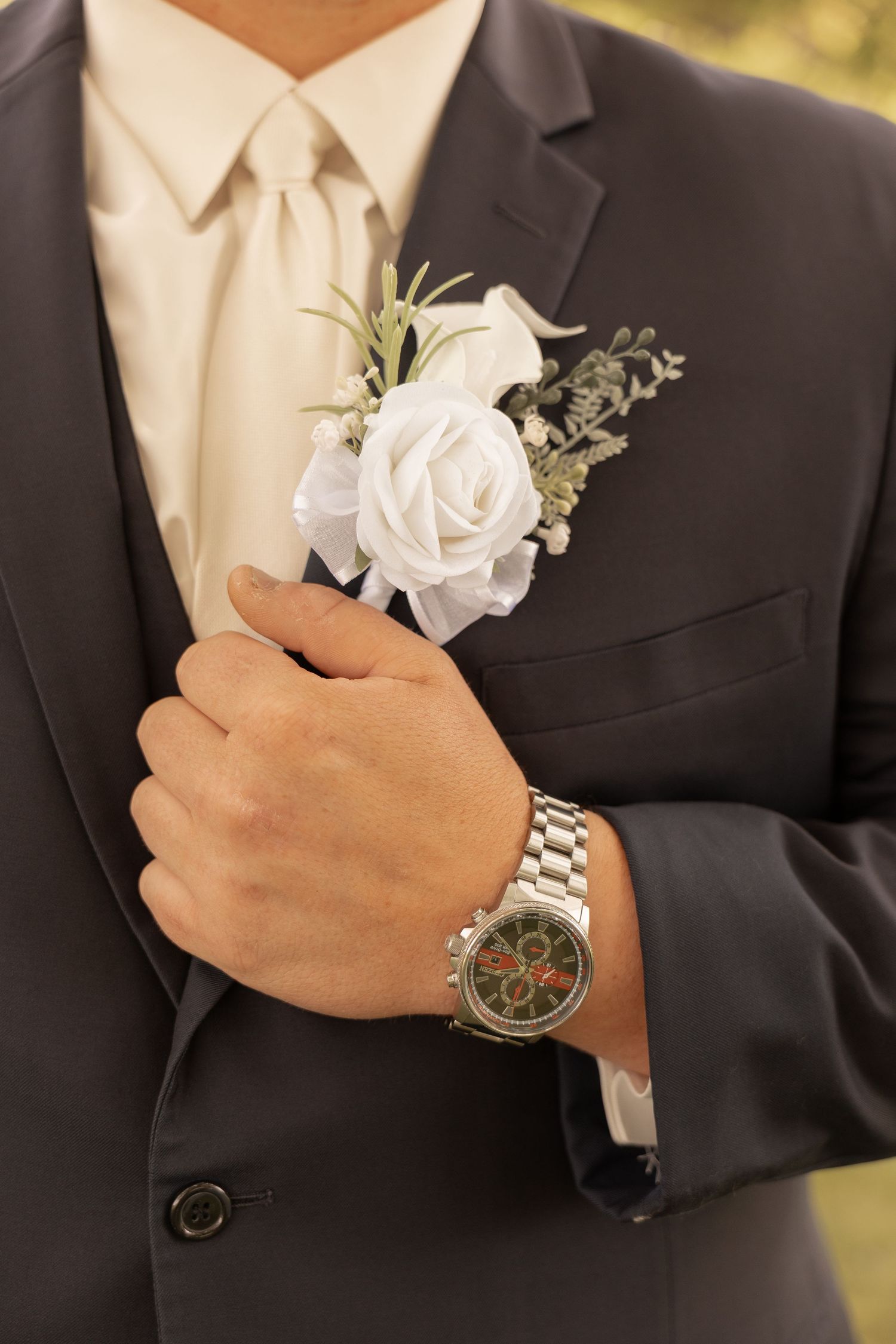Groom adjusting white rose boutonniere on dark suit lapel, wearing silver chronograph watch, with cream tie.