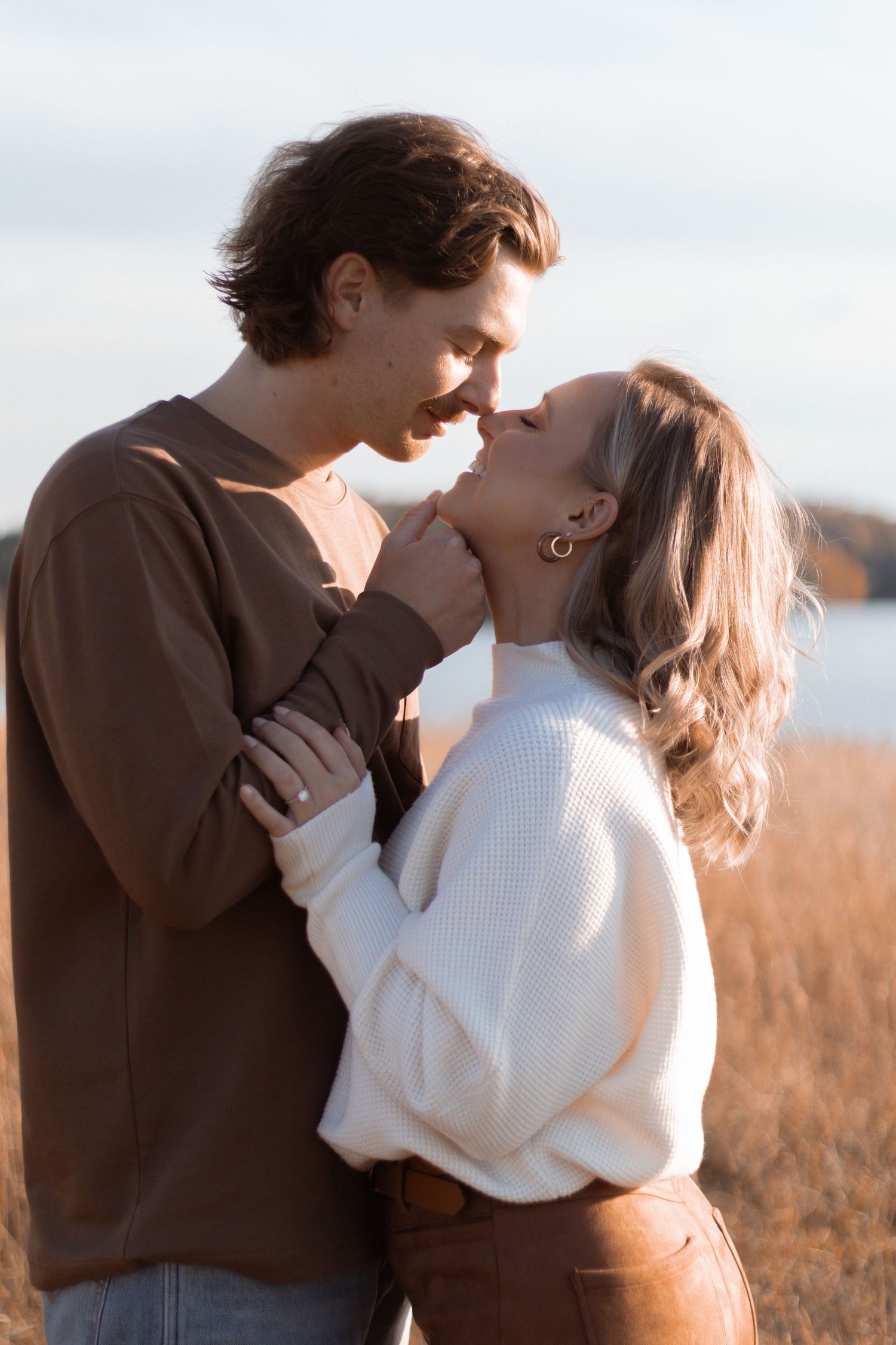 A romantic couple in cozy sweaters share an intimate moment in a sunlit field by the water.