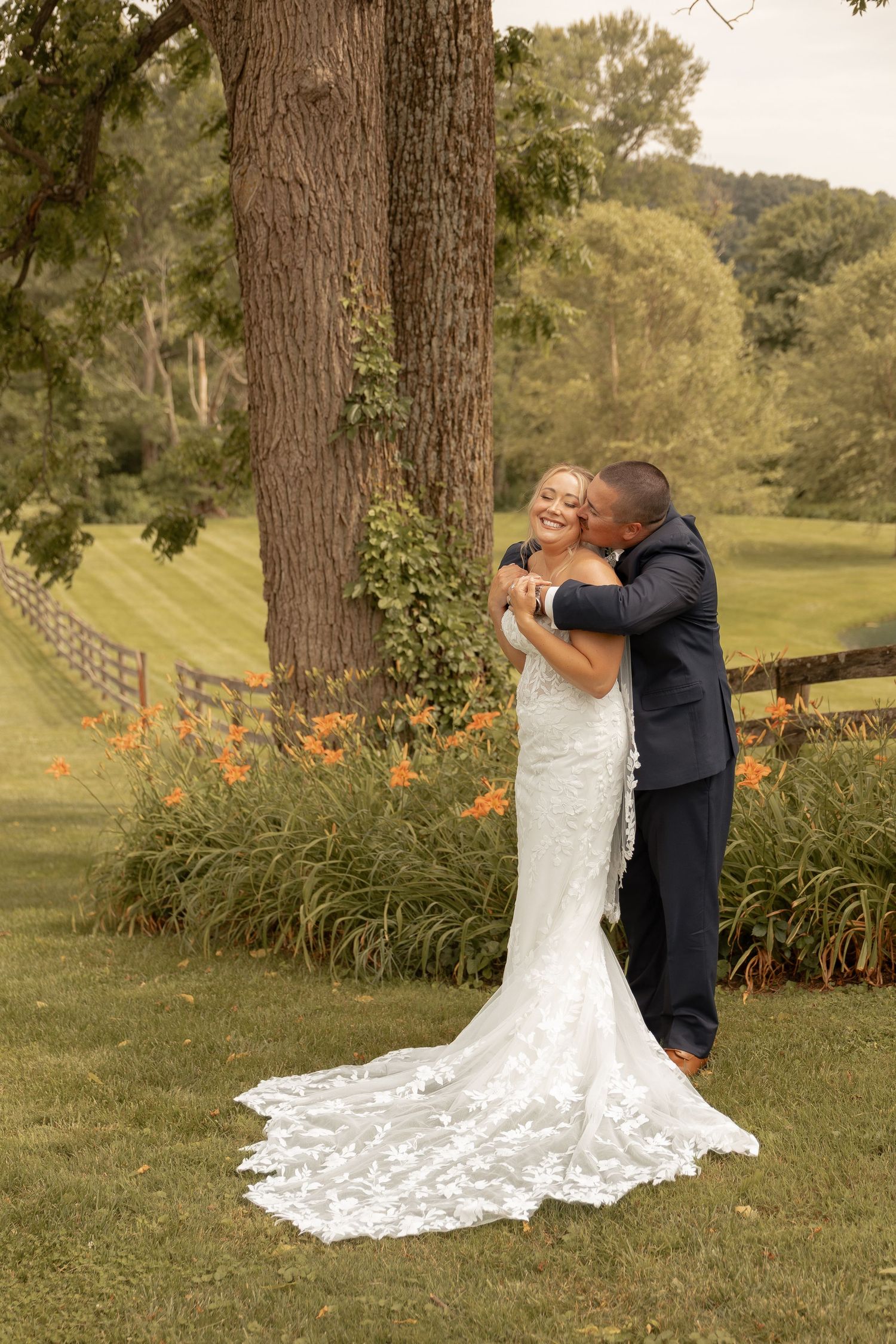 A bride in a lace gown and groom in navy suit embrace joyfully under a large tree at an outdoor wedding venue.