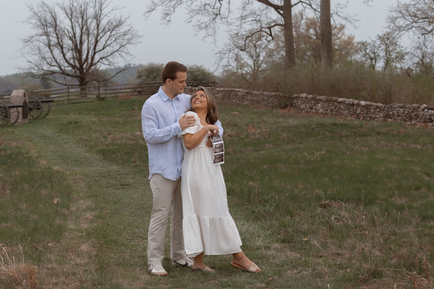 A couple in white attire shares a romantic moment on a grassy hillside with a rustic wooden fence in the background.