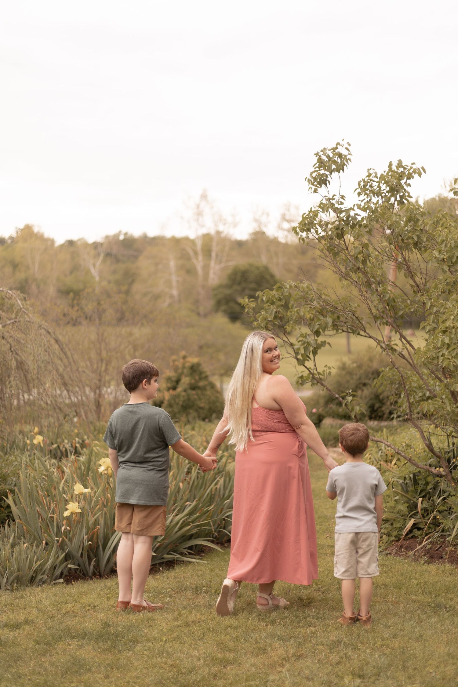 A mother in a pink dress holds hands with her two young sons in a lush green garden setting.