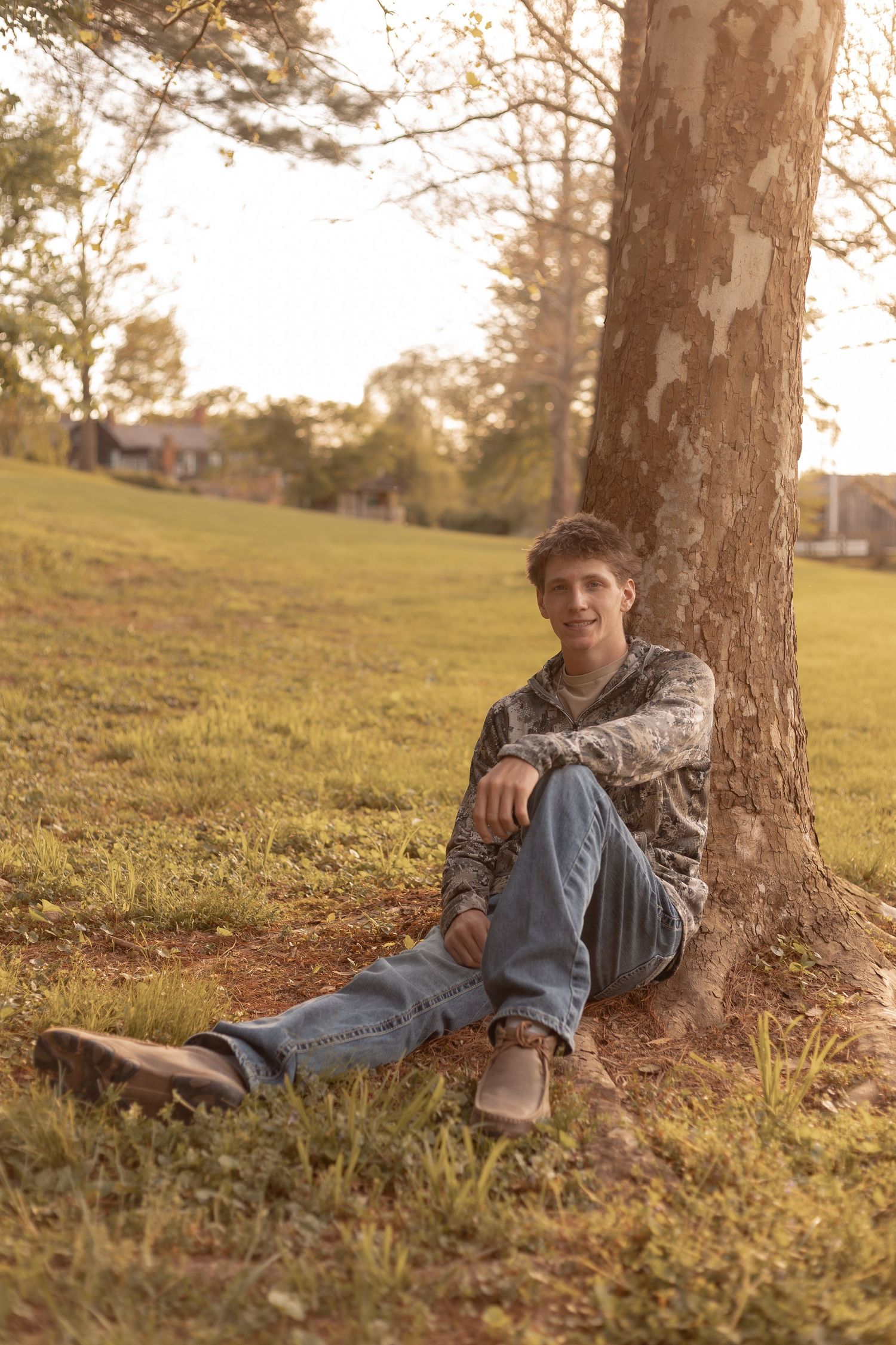 Young man in camo jacket and jeans sitting against a tree in a sunny green park, smiling at camera.