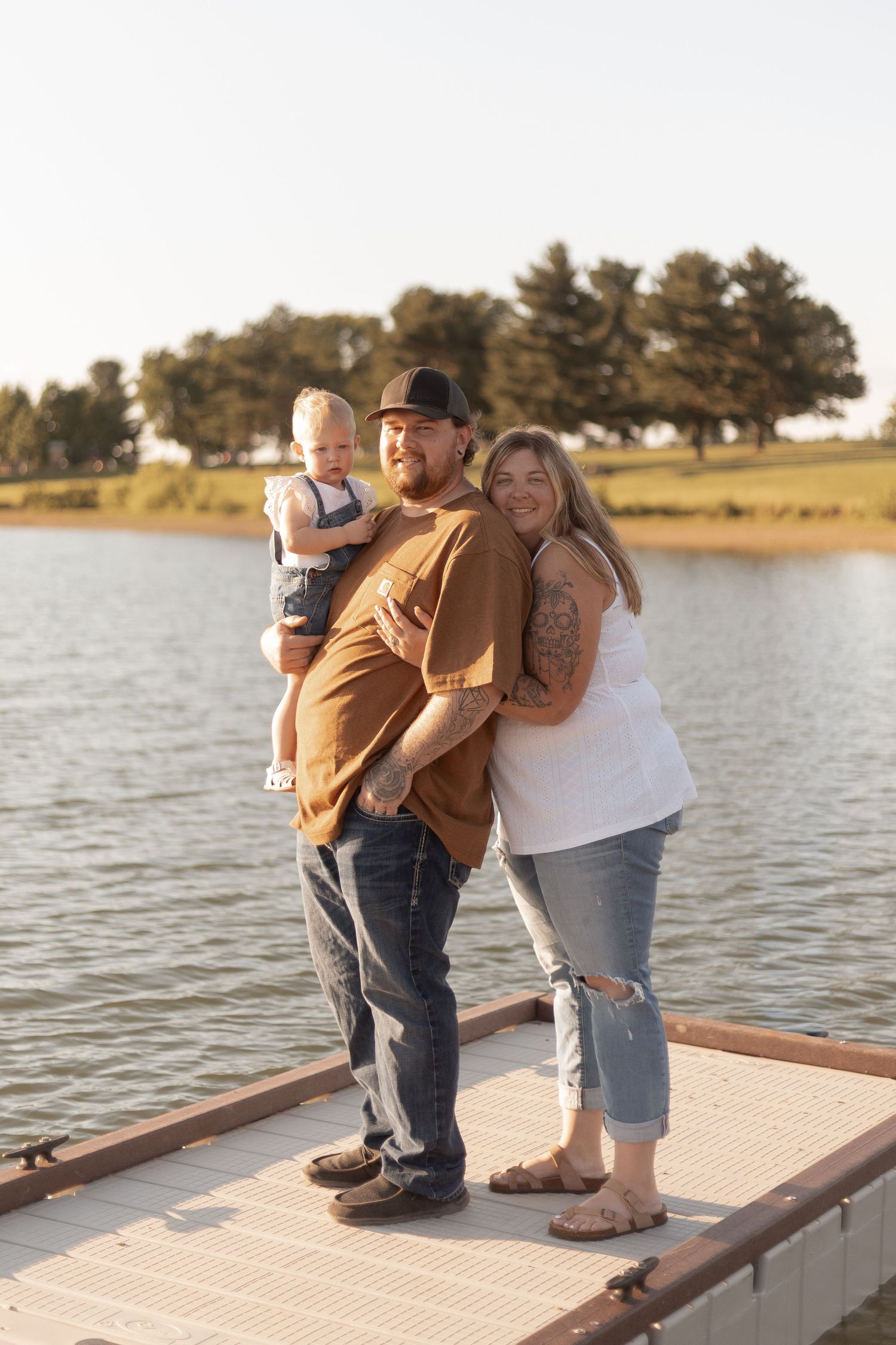 Happy family of three posing on a lakeside dock, parents holding toddler, trees and calm water in background.