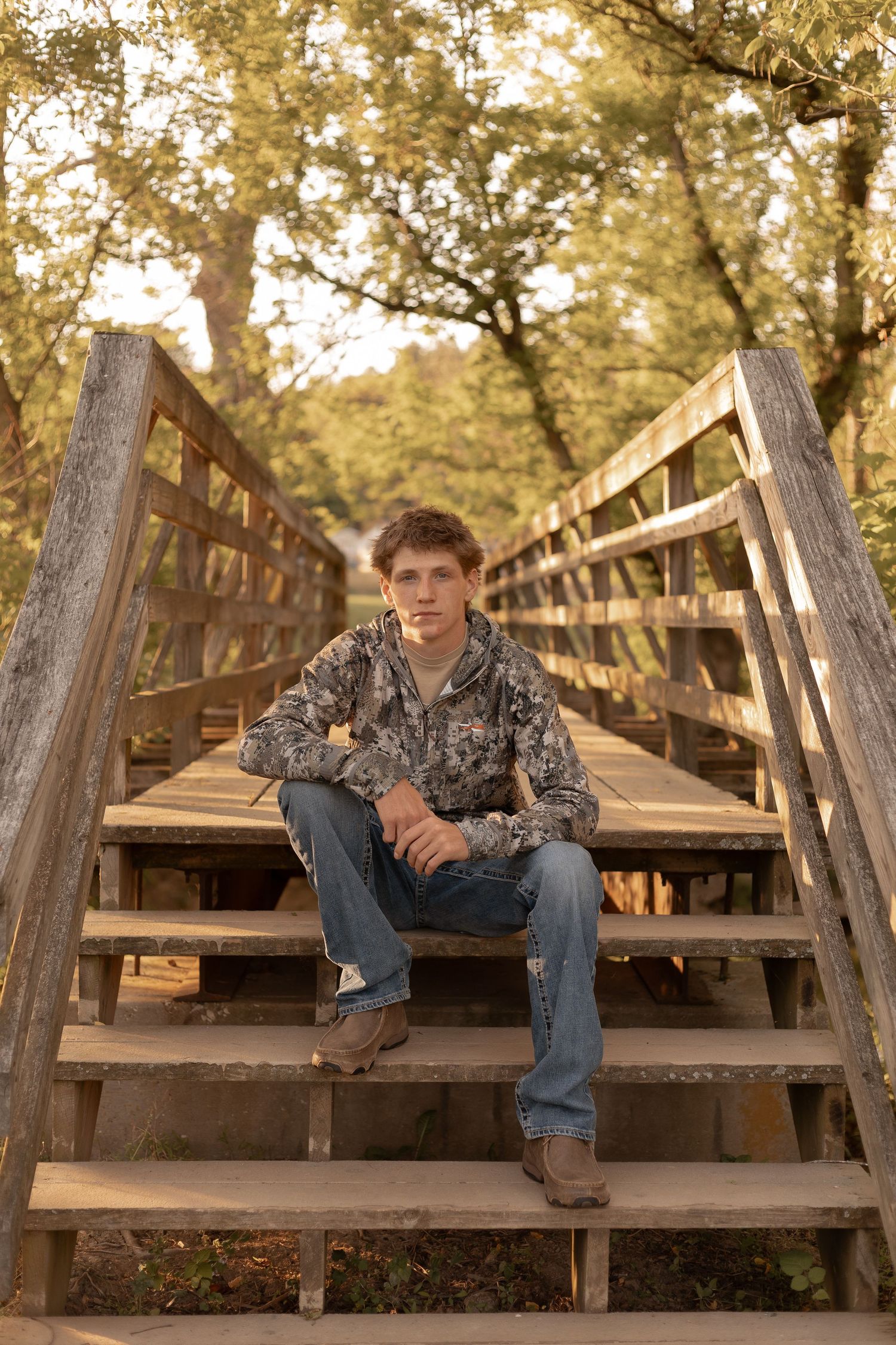 Young man in camo hoodie and jeans sitting on wooden bridge steps surrounded by autumn trees.