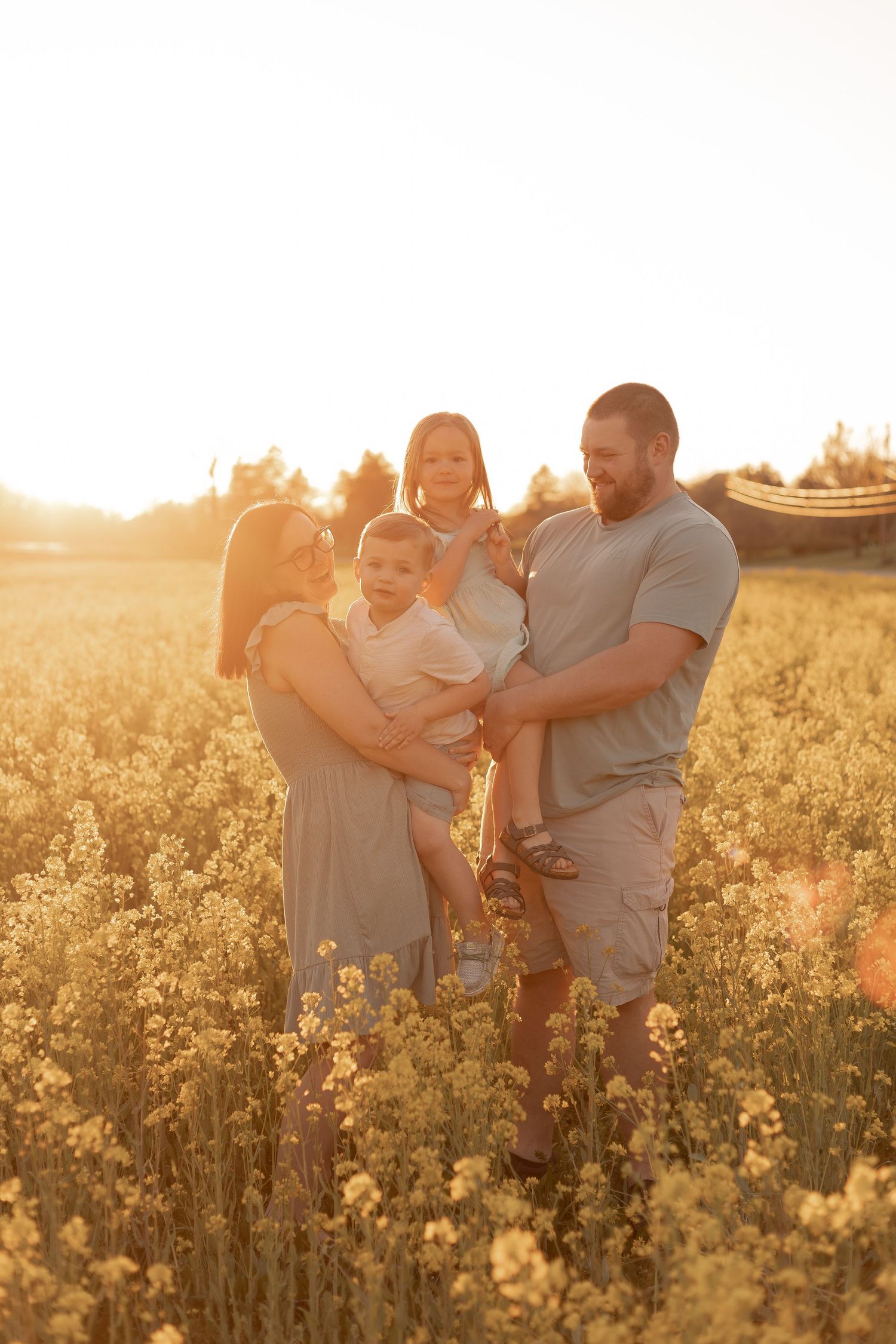 Family of four posing in golden sunlit rapeseed field at sunset, parents holding young children among yellow wildflowers.
