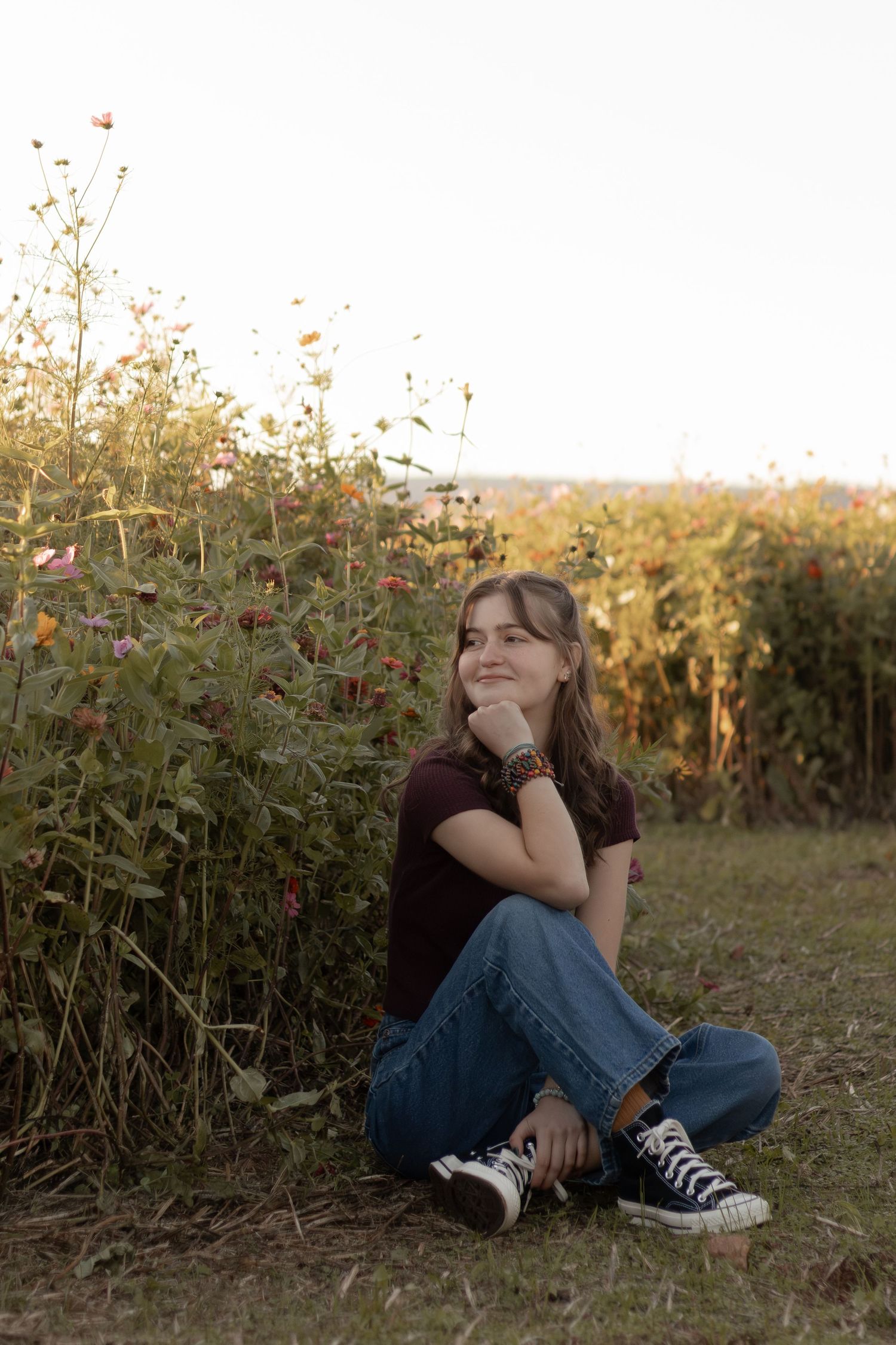 Young girl sitting cross-legged in a wildflower field, wearing jeans and Converse sneakers, smiling warmly at golden hour.