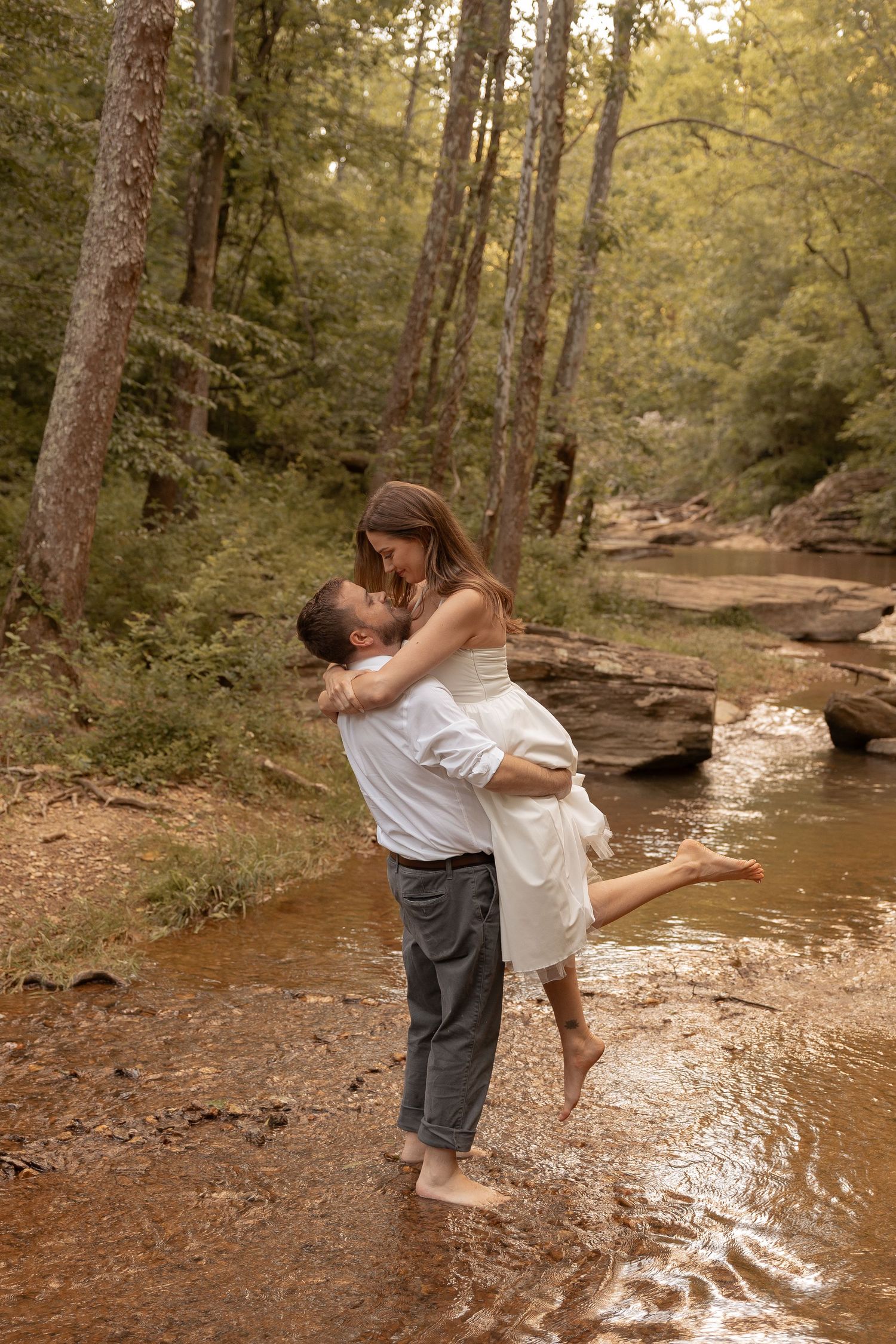 Couple sharing romantic moment in forest creek, man lifting woman in white dress among lush trees and rocky stream.