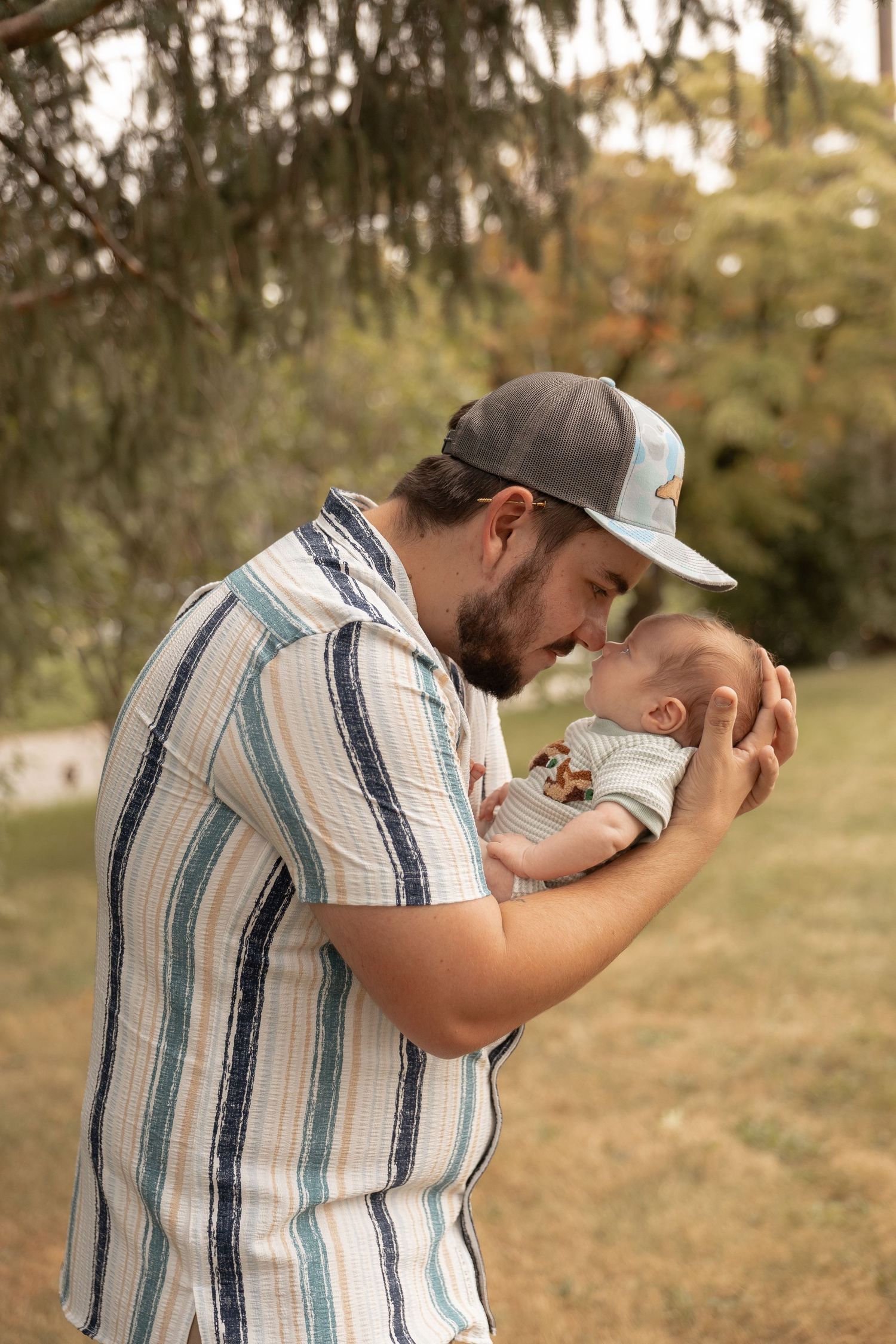 A bearded father in a striped shirt and cap tenderly holds his newborn baby outdoors in a lush green park.