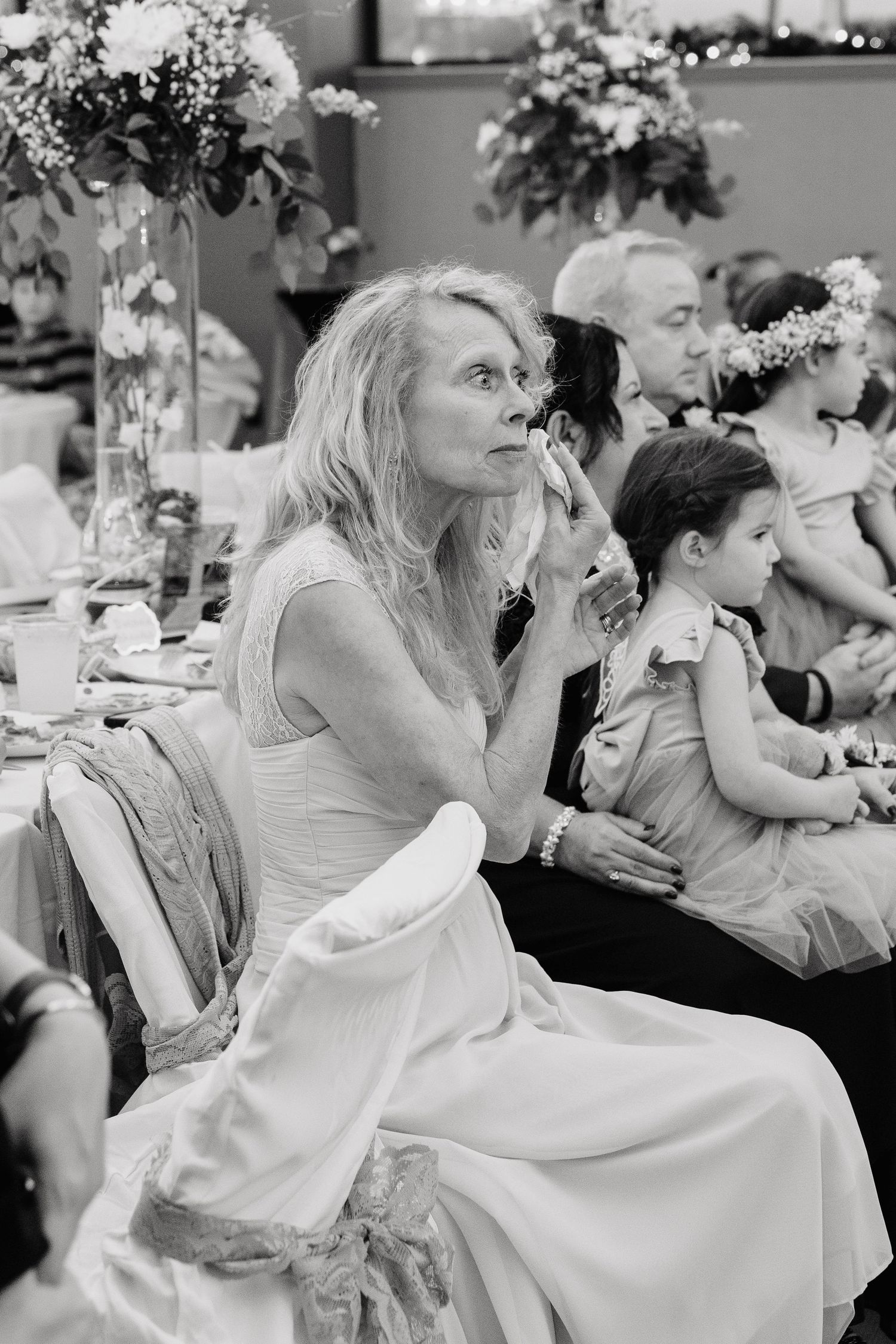 Black and white photo of emotional wedding guests seated at a reception table, woman in white dress wiping tears.