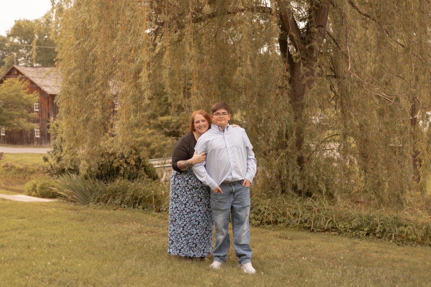 Couple posing together under a weeping willow tree on a green lawn near a rustic building.