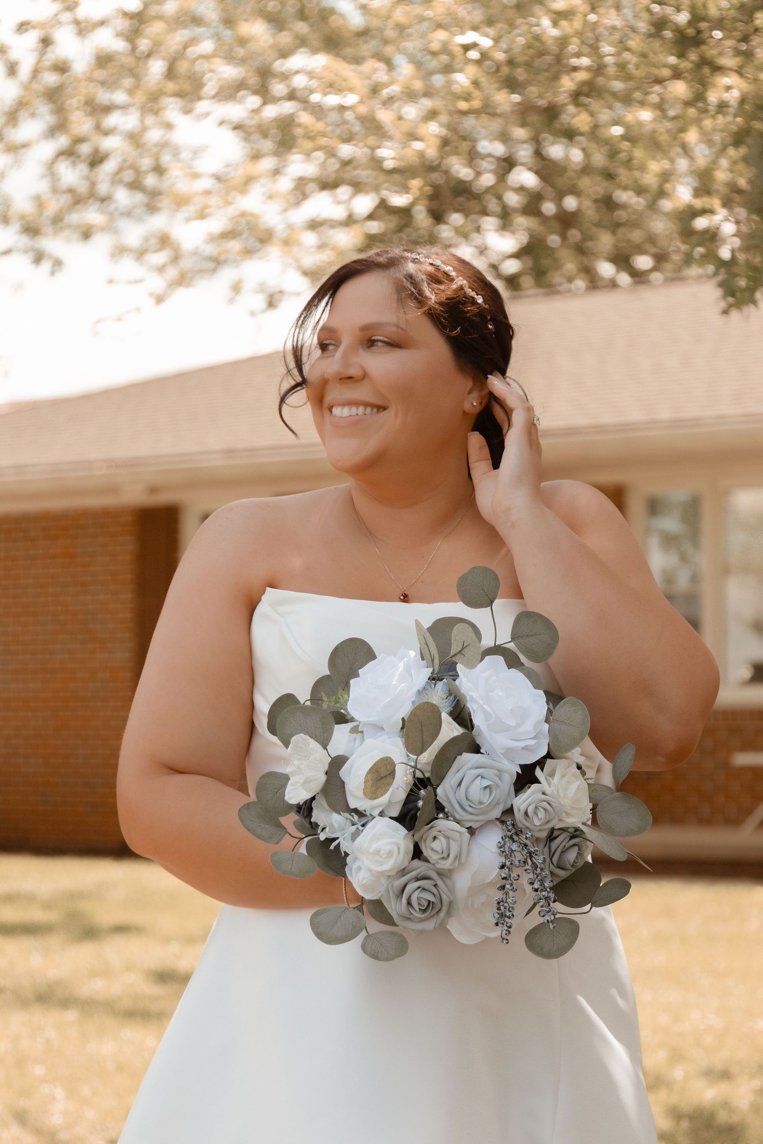 Smiling bride in strapless white gown holding bouquet of white and gray roses with eucalyptus outdoors.