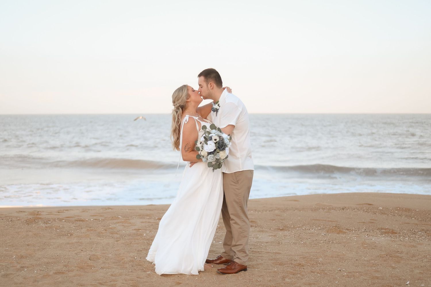 Bride and groom share a romantic kiss on a sandy beach at sunset, bride holding a white floral bouquet.