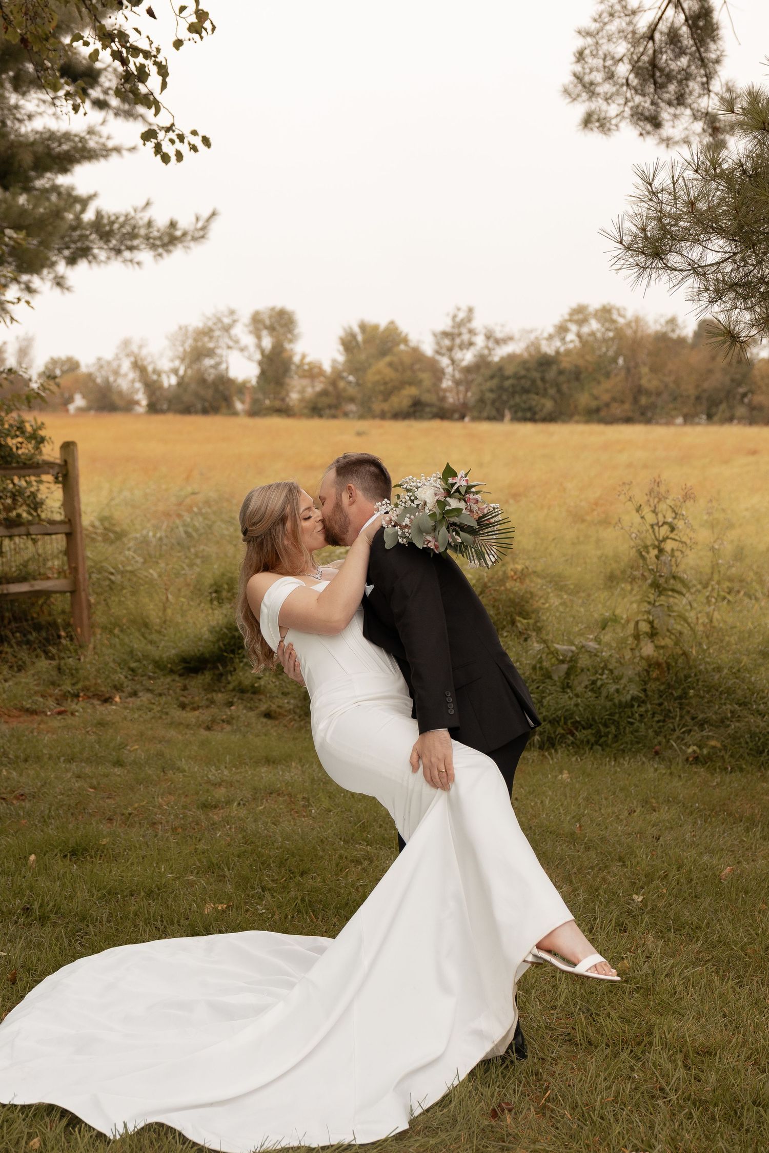 Groom dips bride for a kiss outdoors, she holds a bouquet in a white gown, with a golden field backdrop.