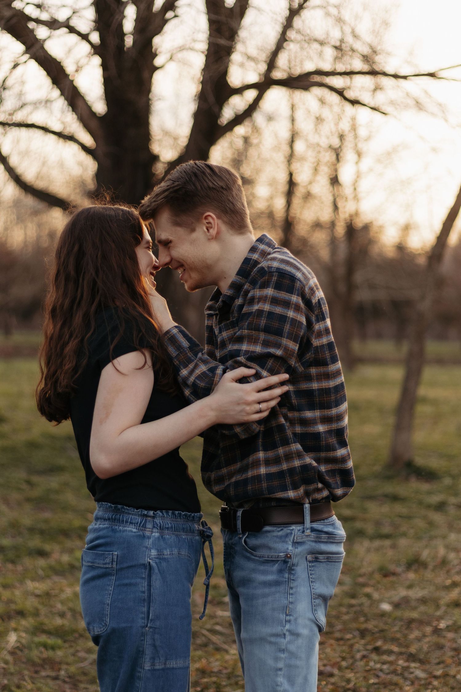 A young couple shares a romantic moment outdoors at sunset, smiling and embracing near bare trees in casual denim attire.