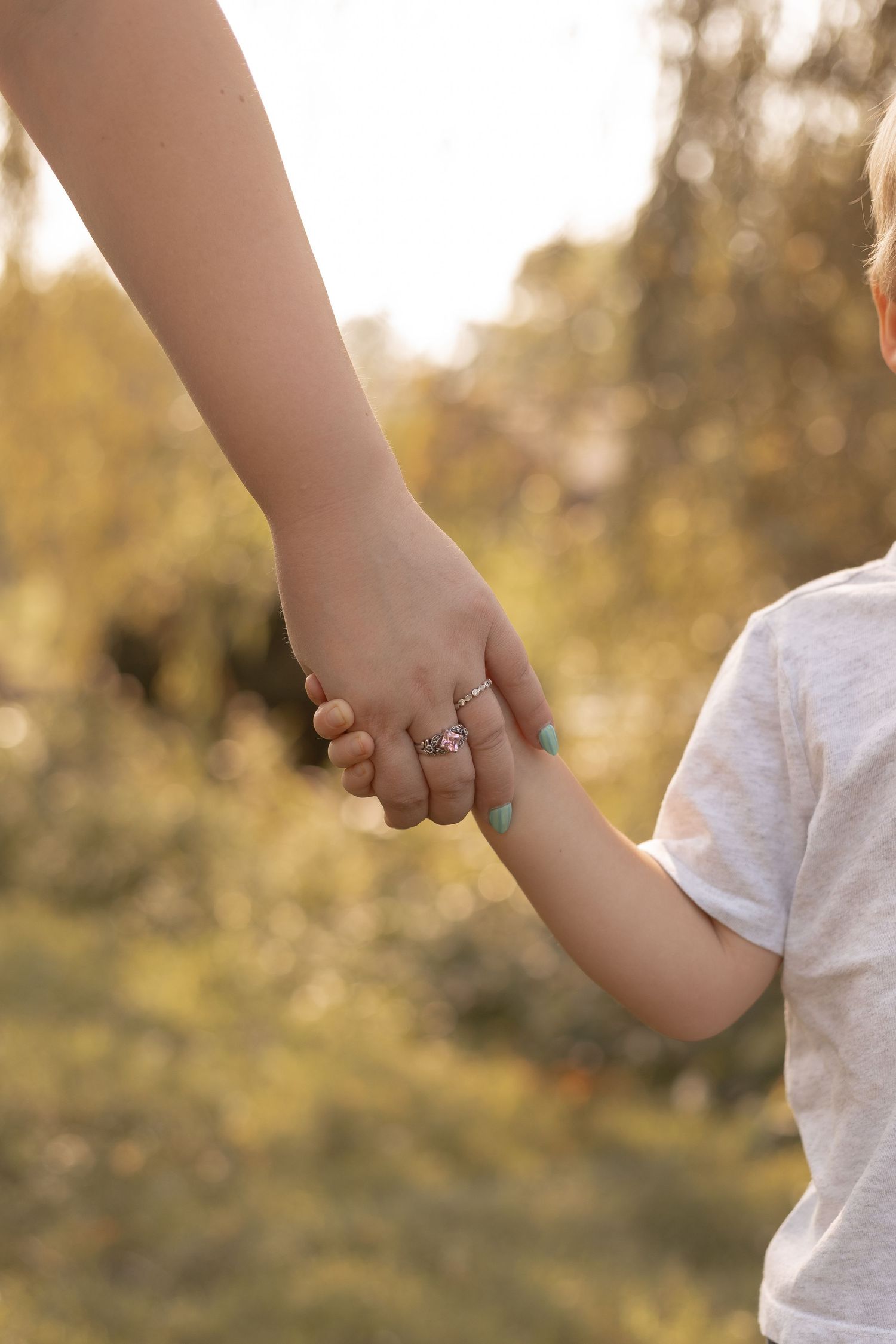 An adult and child holding hands outdoors in a sunlit park, showcasing a silver ring and teal nail polish.