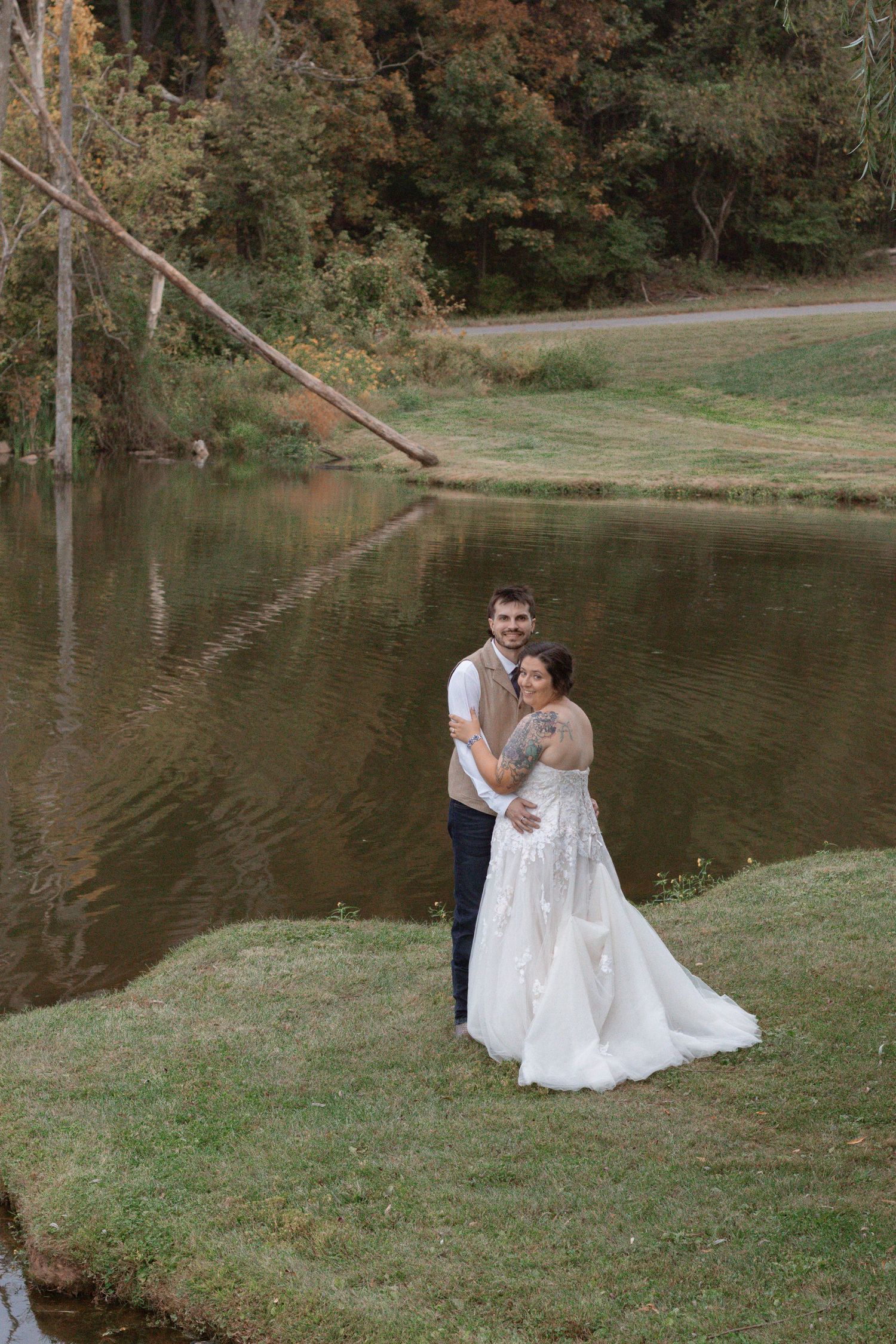 A couple shares a romantic moment by a tranquil pond surrounded by autumn trees and grass.