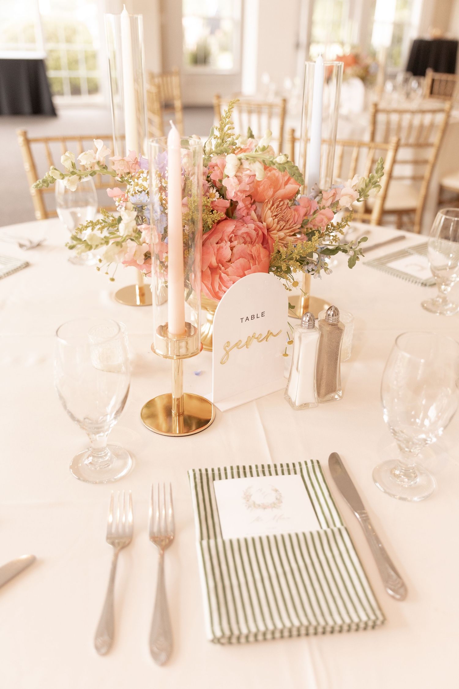 Elegant wedding reception table with pink peony centerpiece, gold candlesticks, striped napkin, and heart-shaped table number card.