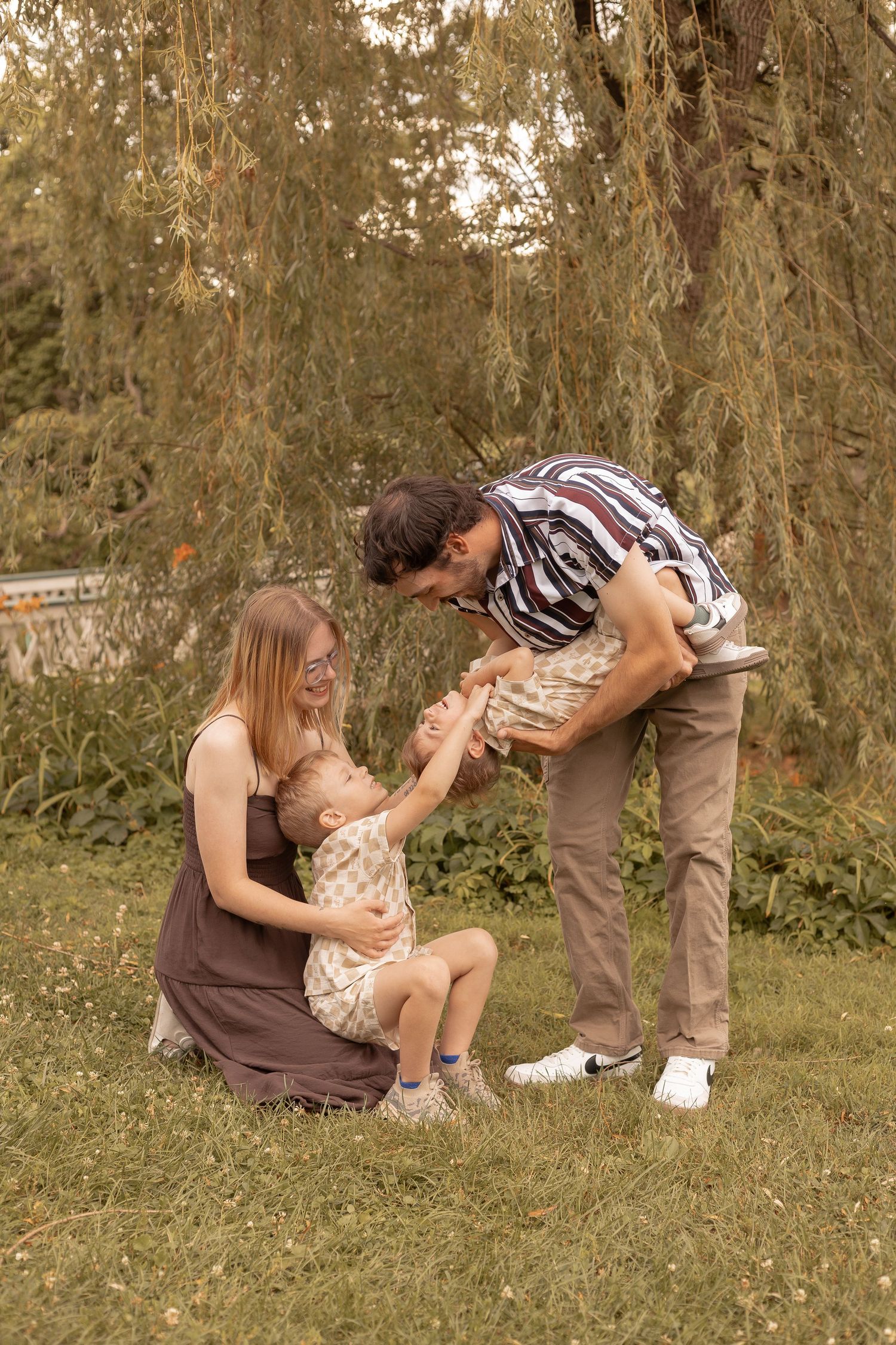 Happy family with two young children playing together on green grass under a willow tree outdoors.