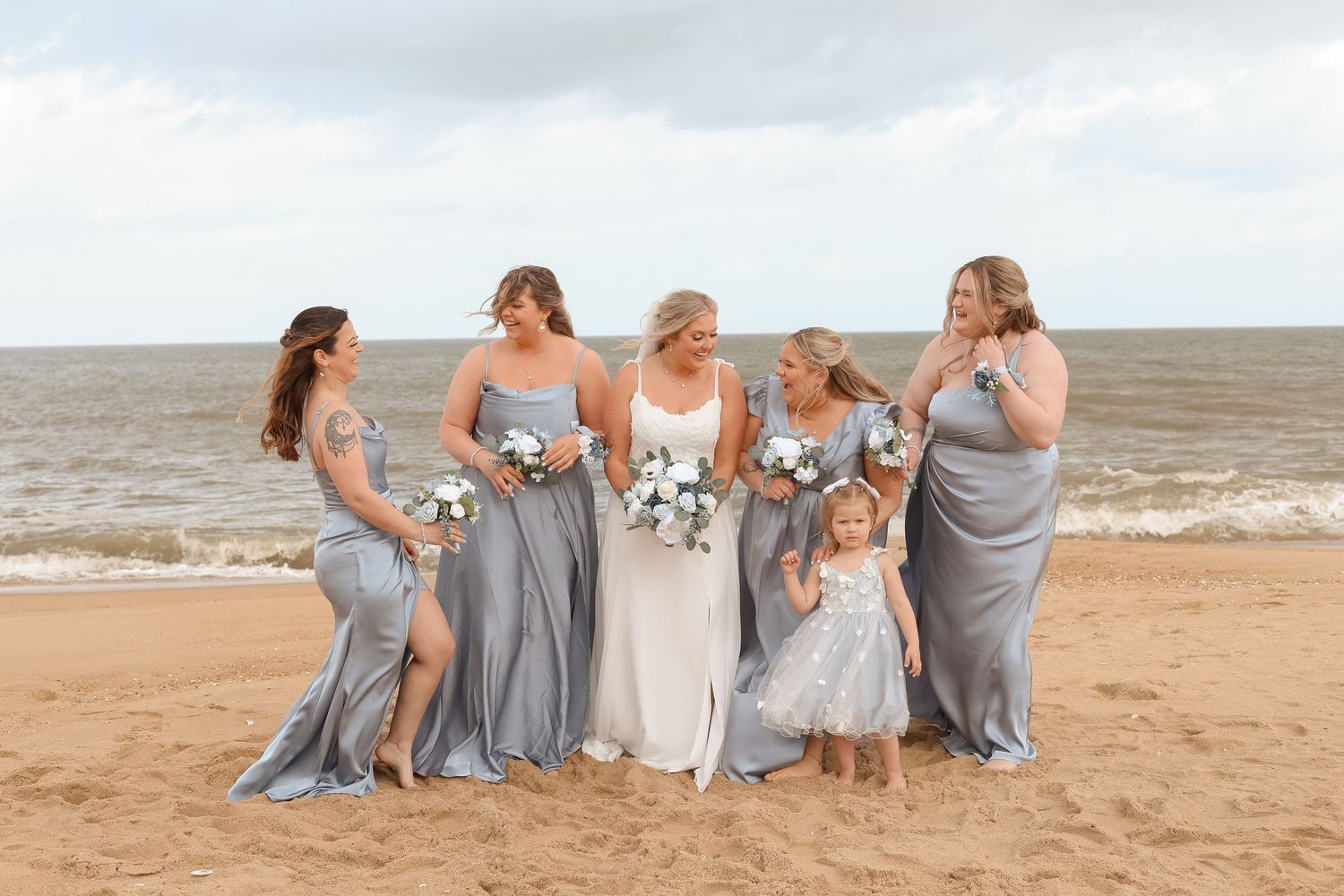 Bride in white gown with bridesmaids in silver dresses and flower girl on sandy beach with ocean backdrop.