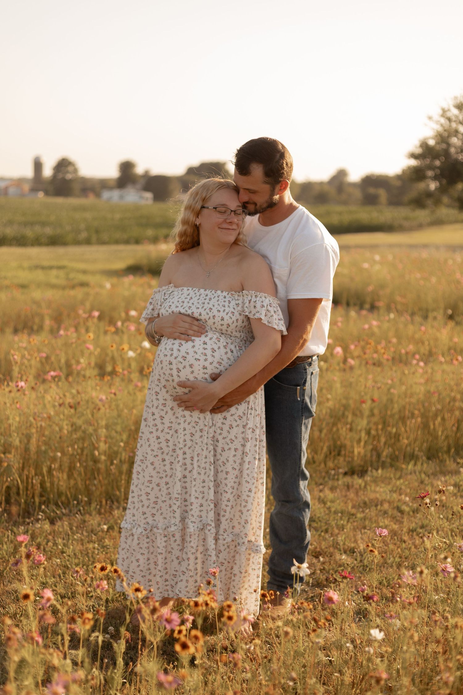 Couple embracing in a sunny wildflower field, pregnant woman in floral dress with partner in white tee and jeans.