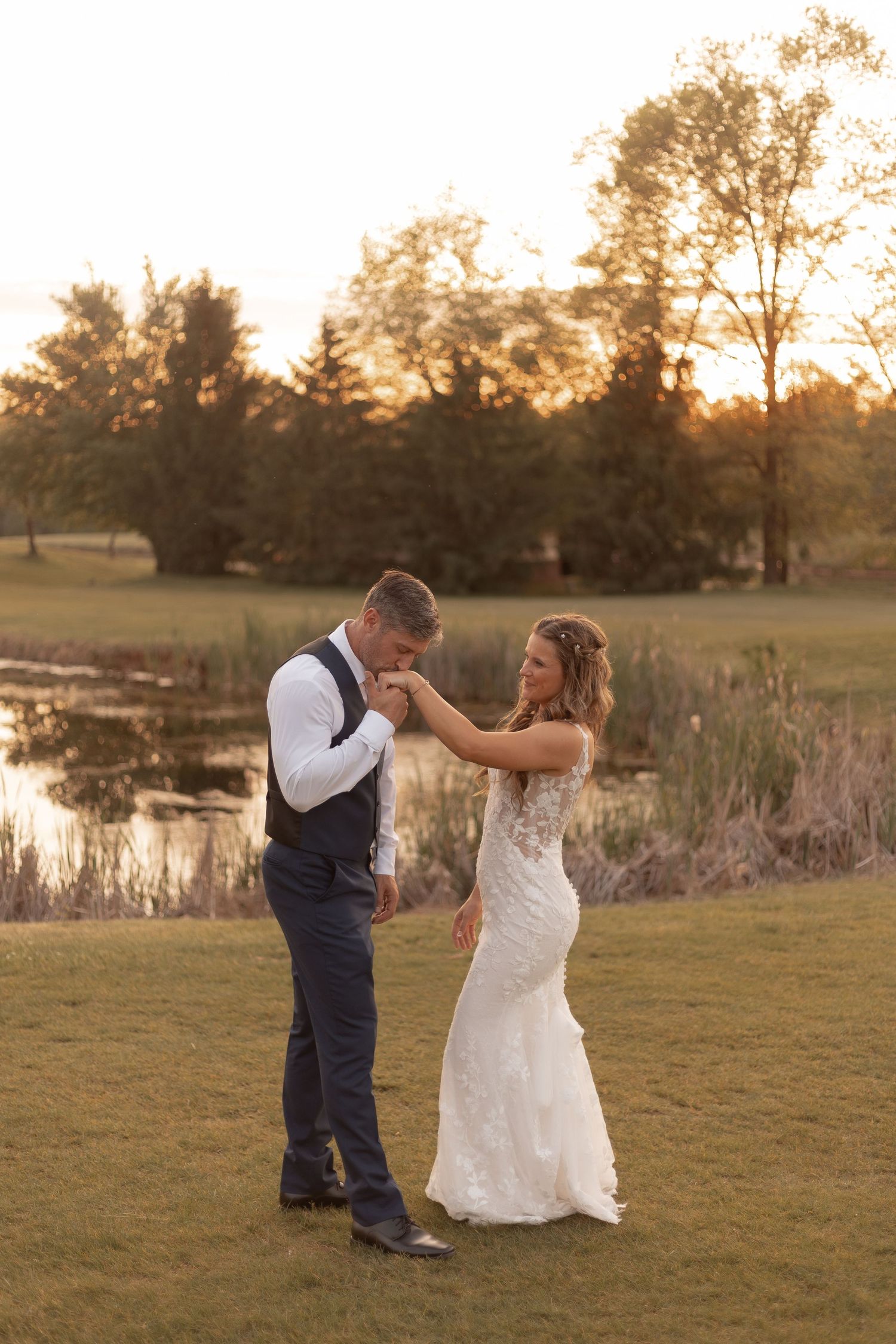 Bride and groom share a romantic first dance on a lush green lawn at golden hour near a serene pond.