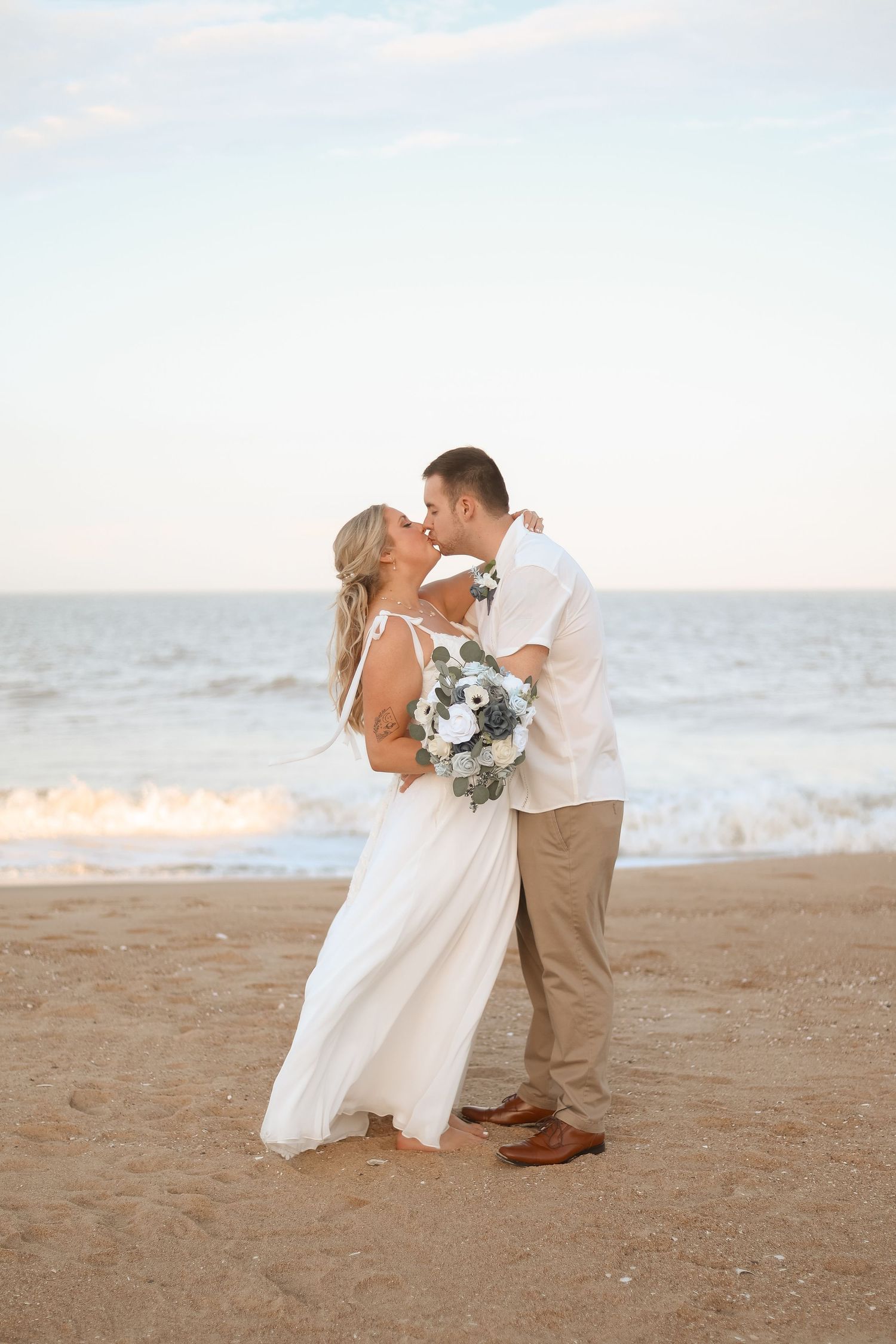Bride and groom kissing on sandy beach at sunset, bride holding white floral bouquet, ocean waves in background.