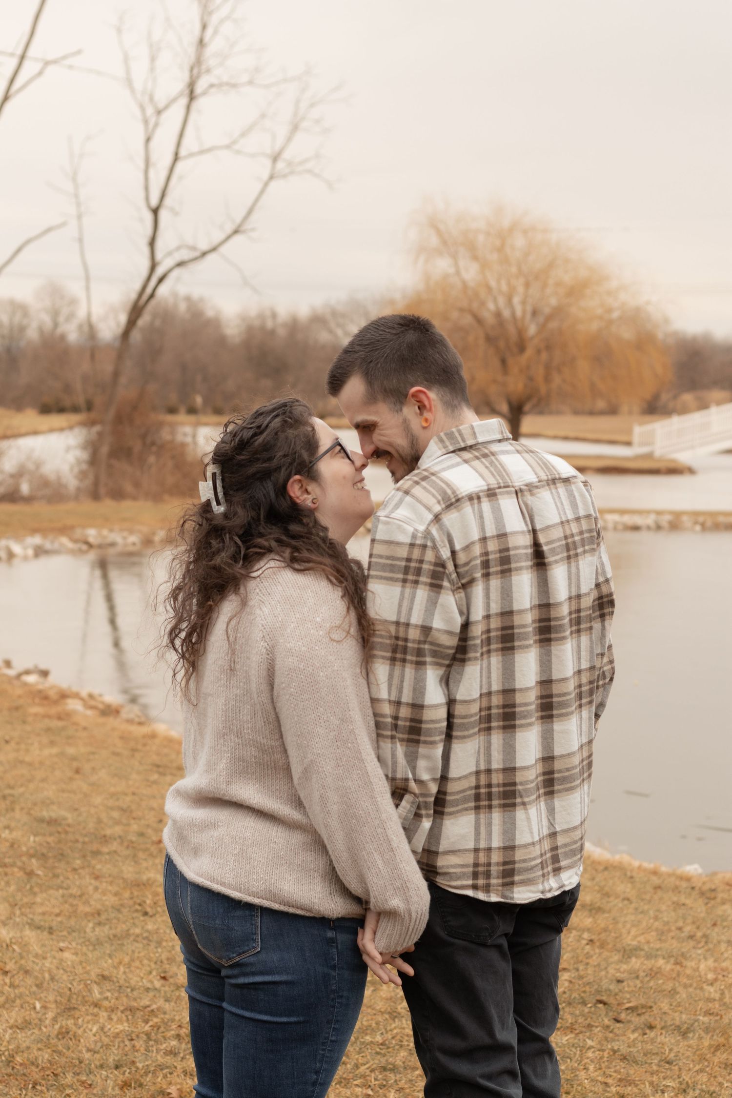 Couple holding hands and sharing a romantic moment by a serene pond with bare winter trees in the background.