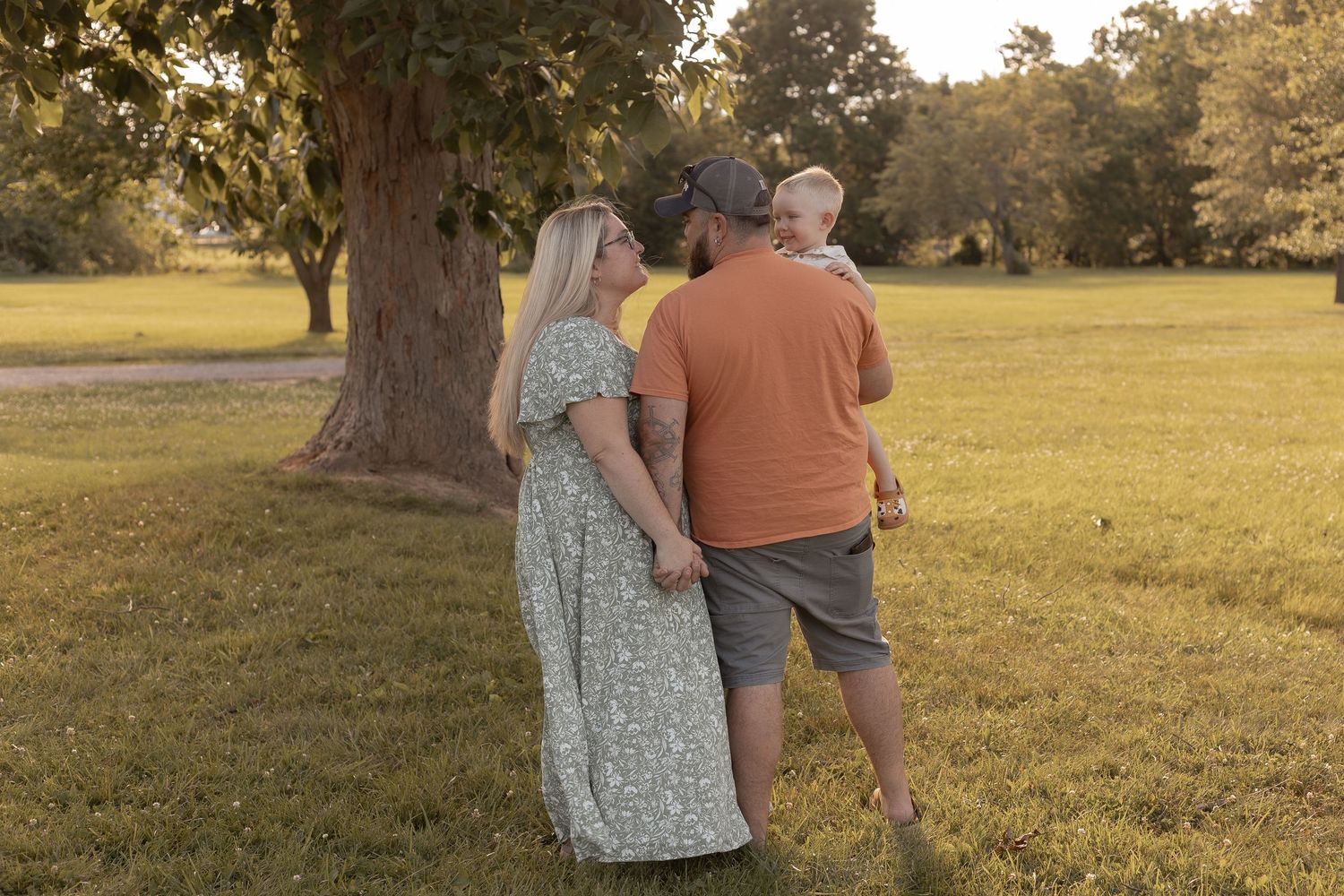 Family walking in a sunny park, parents holding hands while father carries a baby, surrounded by lush green grass and trees.