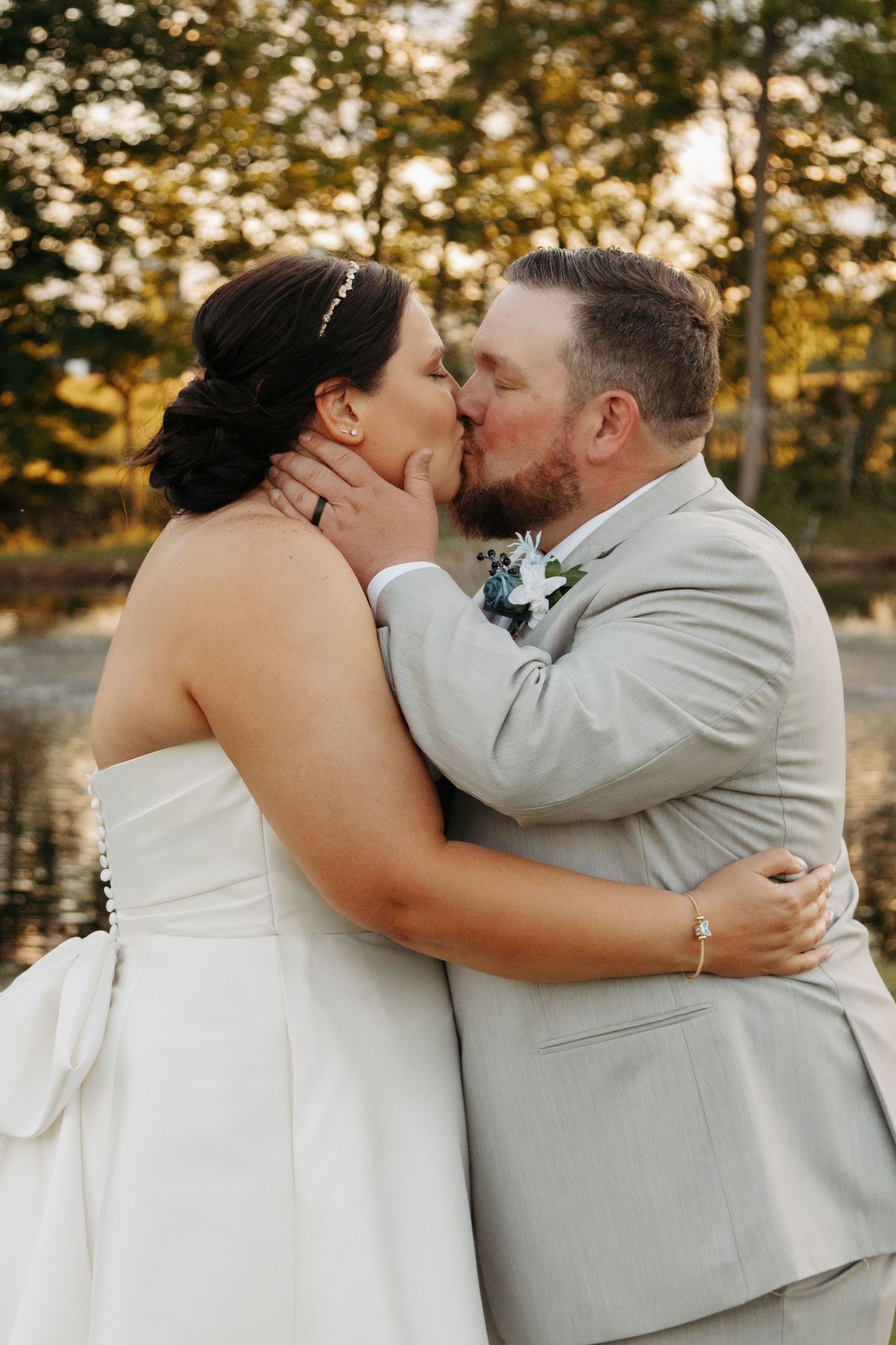 Bride and groom share a romantic first kiss outdoors near a pond, surrounded by autumn trees, groom in gray suit.