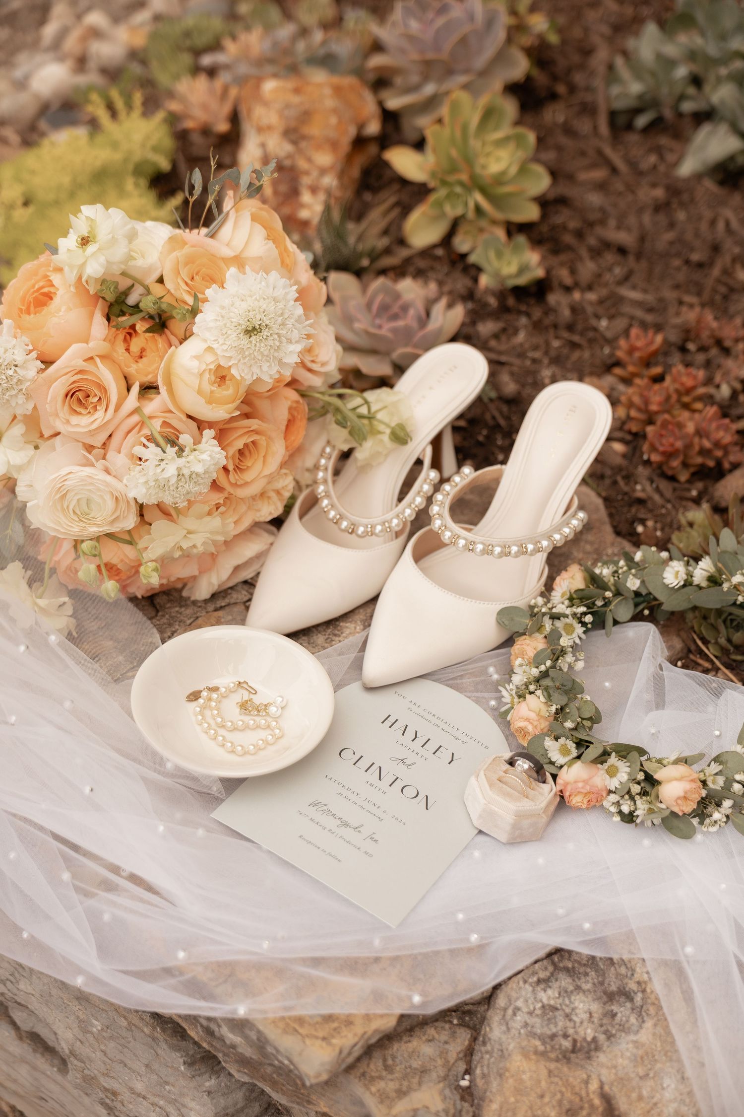 Elegant bridal flat lay with pearl mules, peach rose bouquet, floral crown, wedding invitation, and ring dish on white veil.