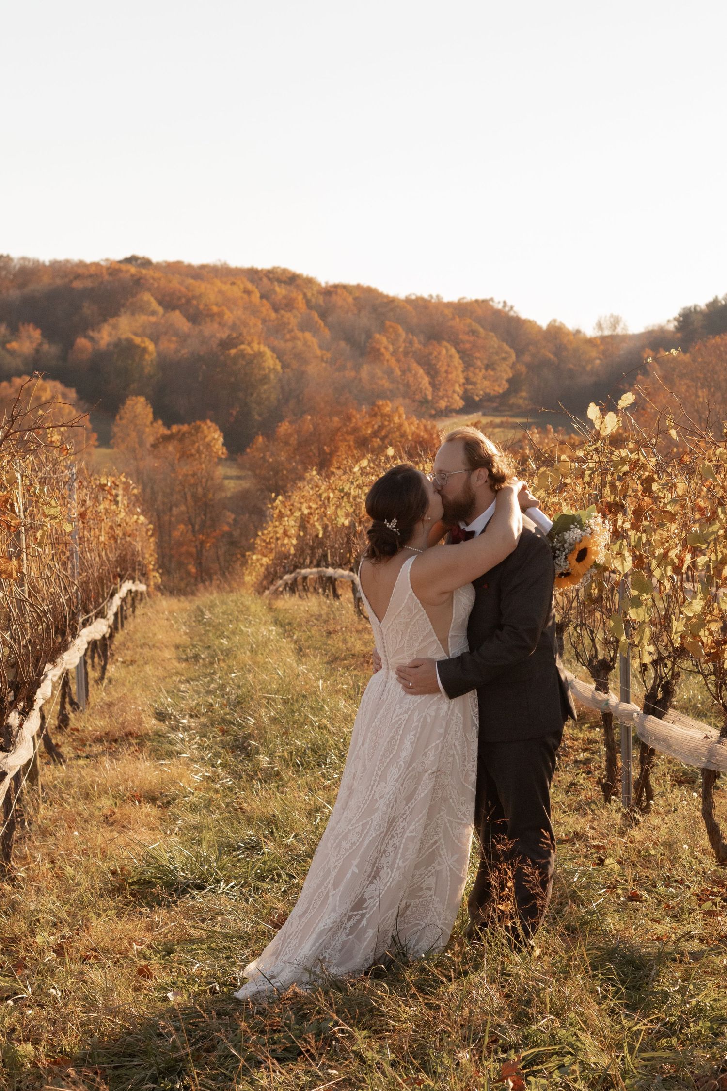 Bride and groom embrace in a sunlit autumn vineyard, surrounded by golden foliage and rolling hills.