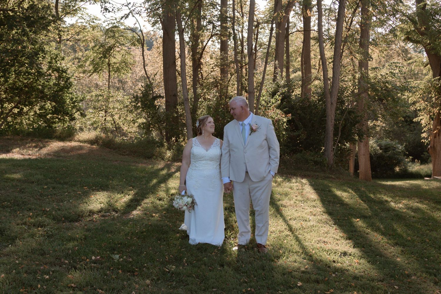 A couple walks together in a sun-dappled wooded area wearing white and light colored clothing.