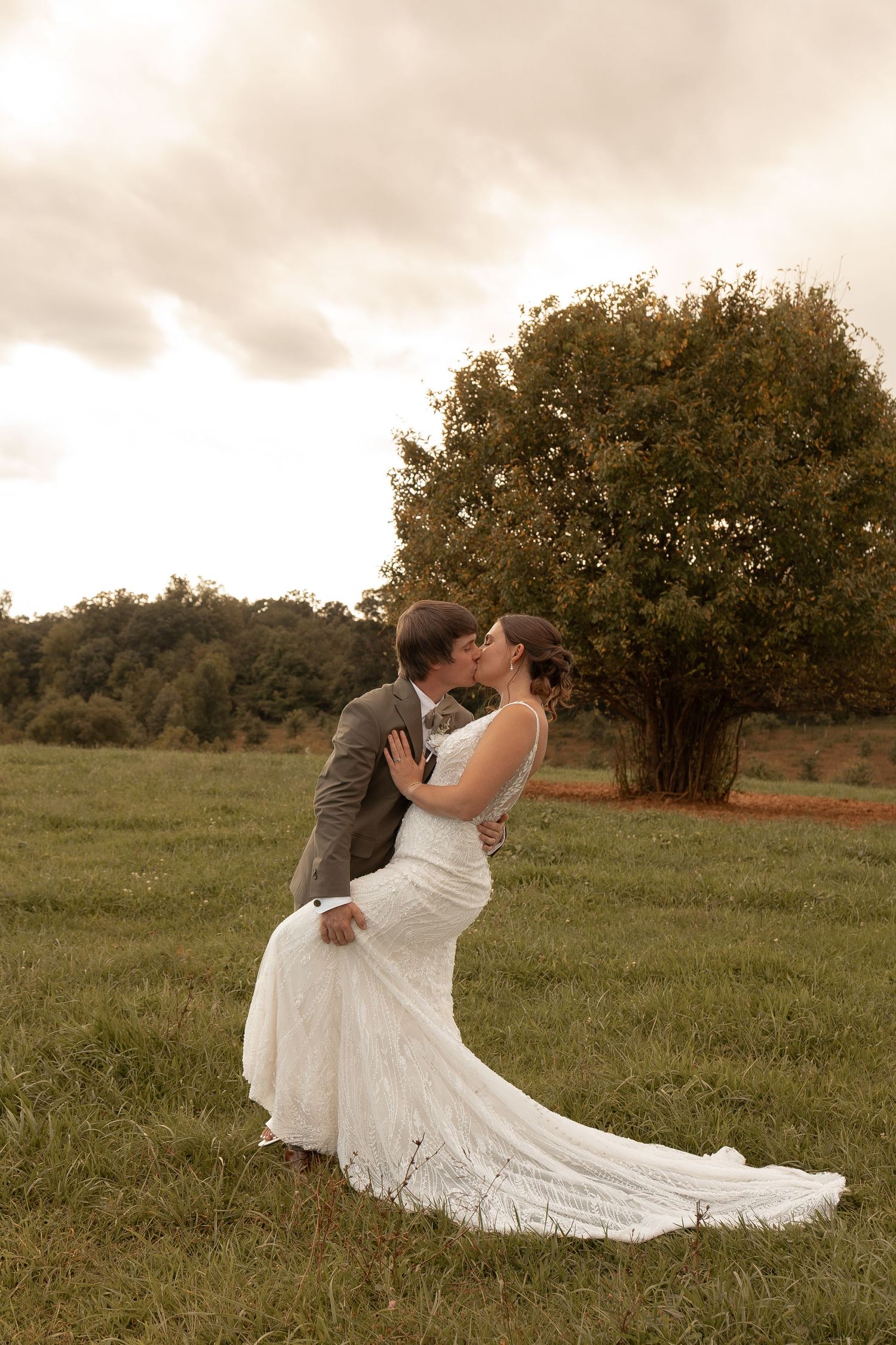 Bride and groom share a romantic kiss in a lush green field with a large tree and cloudy sky backdrop.
