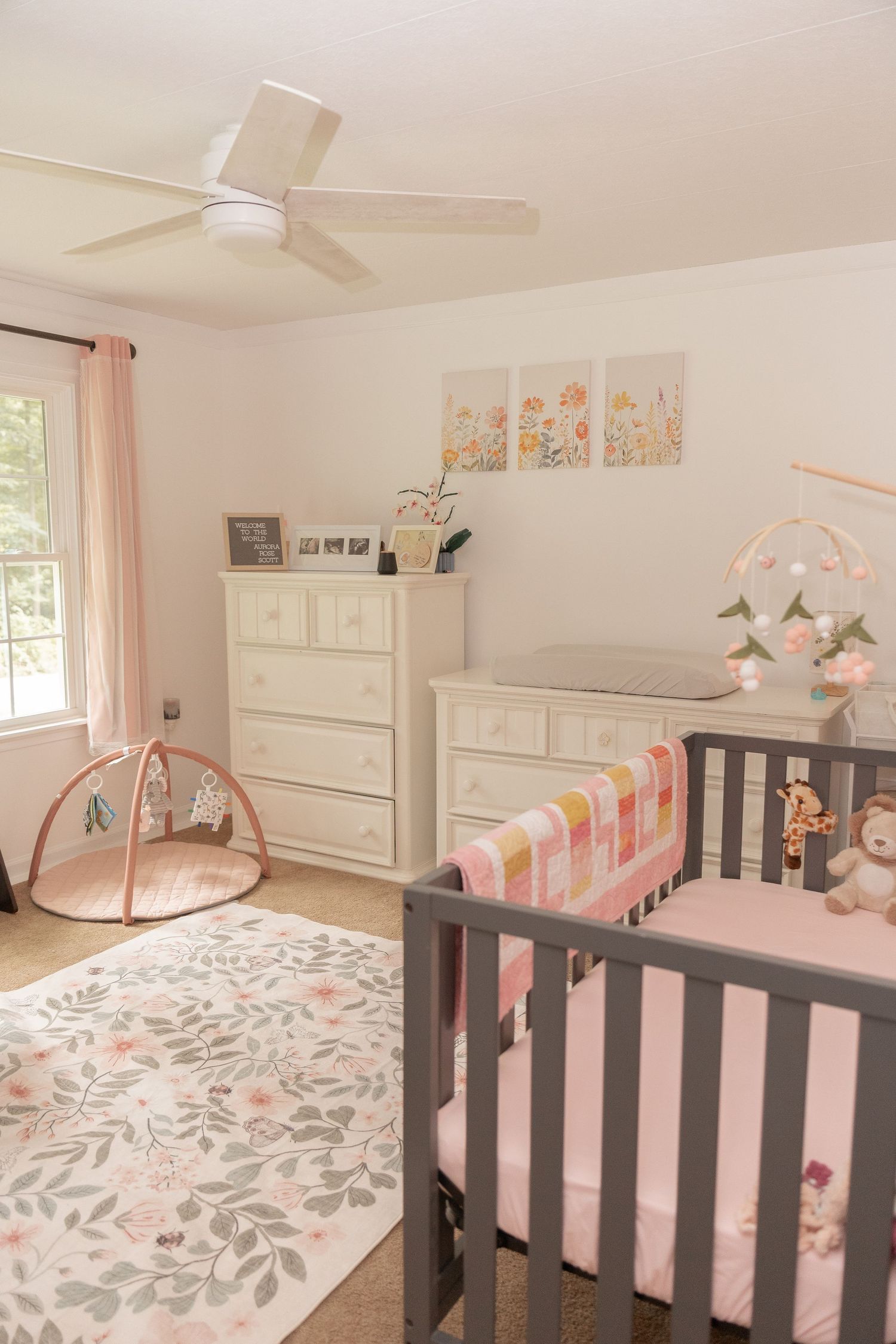 Soft pink nursery with gray crib, white dresser, floral rug, ceiling fan, and colorful wall art for a baby girl.
